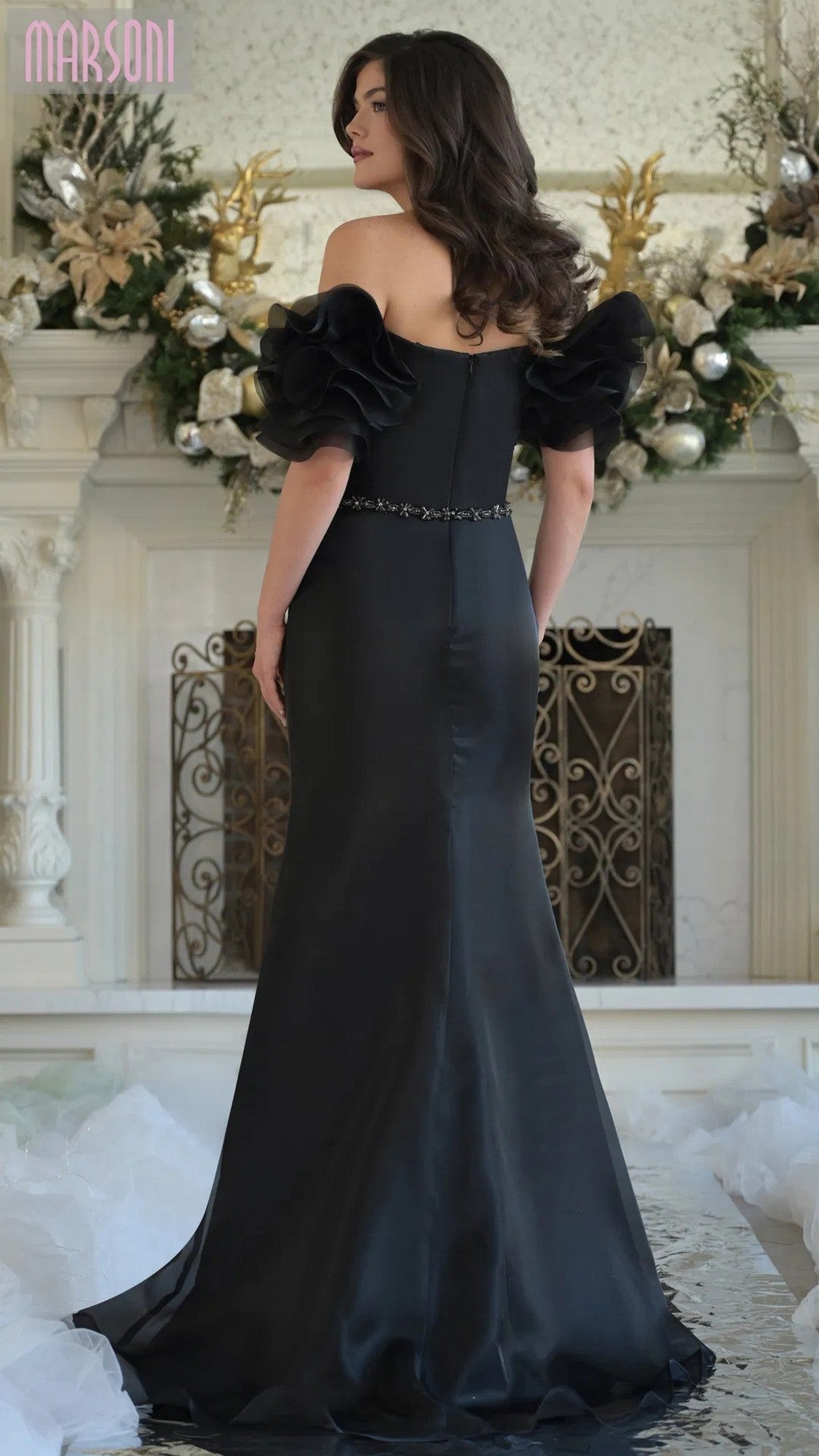 A woman in the Marsoni MV1353 long formal dress with off-shoulder ruffled sleeves stands elegantly before a decorated white fireplace.
