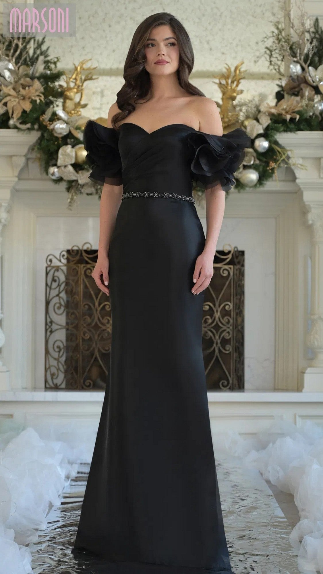 A woman poses gracefully indoors in a black Long Formal Dress: Marsoni MV1353, featuring ruffled off-shoulder sleeves and a fitted waist, standing in front of a decorated fireplace.