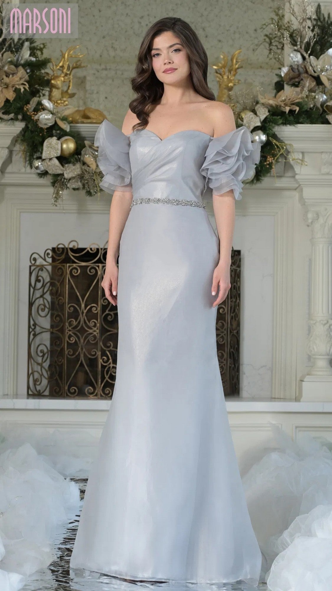 A woman stands indoors by a decorated fireplace, wearing the Marsoni MV1353 long formal dress in oyster, featuring ruffled off-the-shoulder sleeves and a jeweled waist belt.