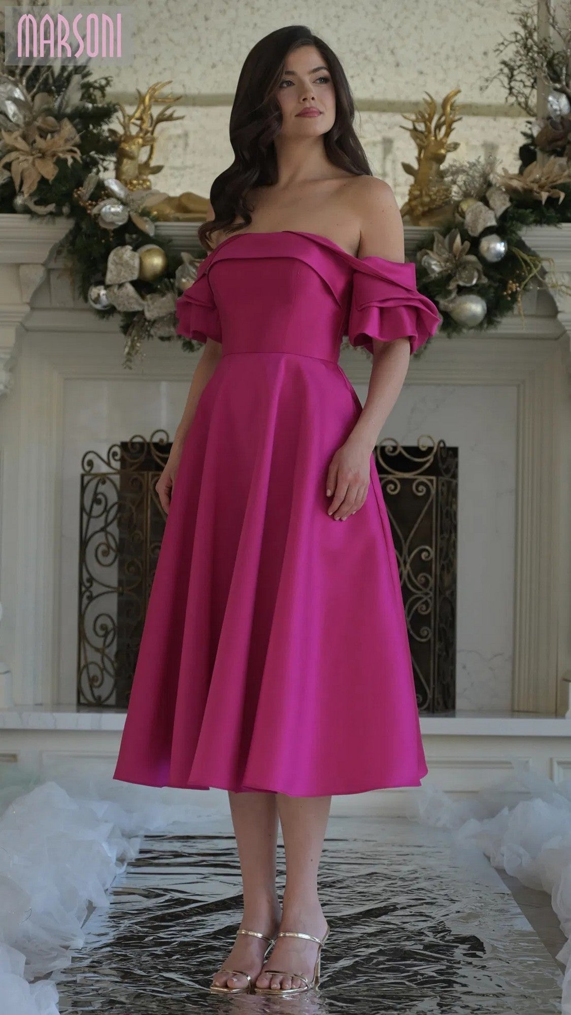 A woman stands indoors wearing the Marsoni MV1369 knee-length semi-formal dress in fuchsia with off-shoulder short sleeves and ruffled details, paired with silver sandals; a decorated mantel is in the background.
