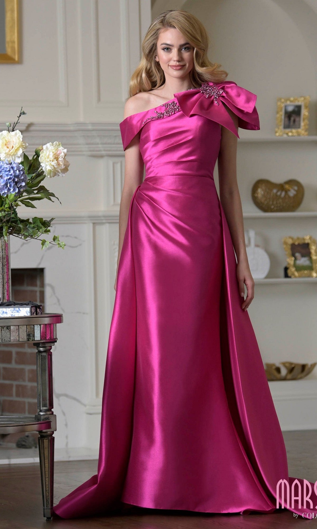 A woman stands indoors wearing the Long Formal Dress: Marsoni MV1408 in fuchsia—a floor-length, off-shoulder gown with a bow accent and floral embellishments on the bodice.