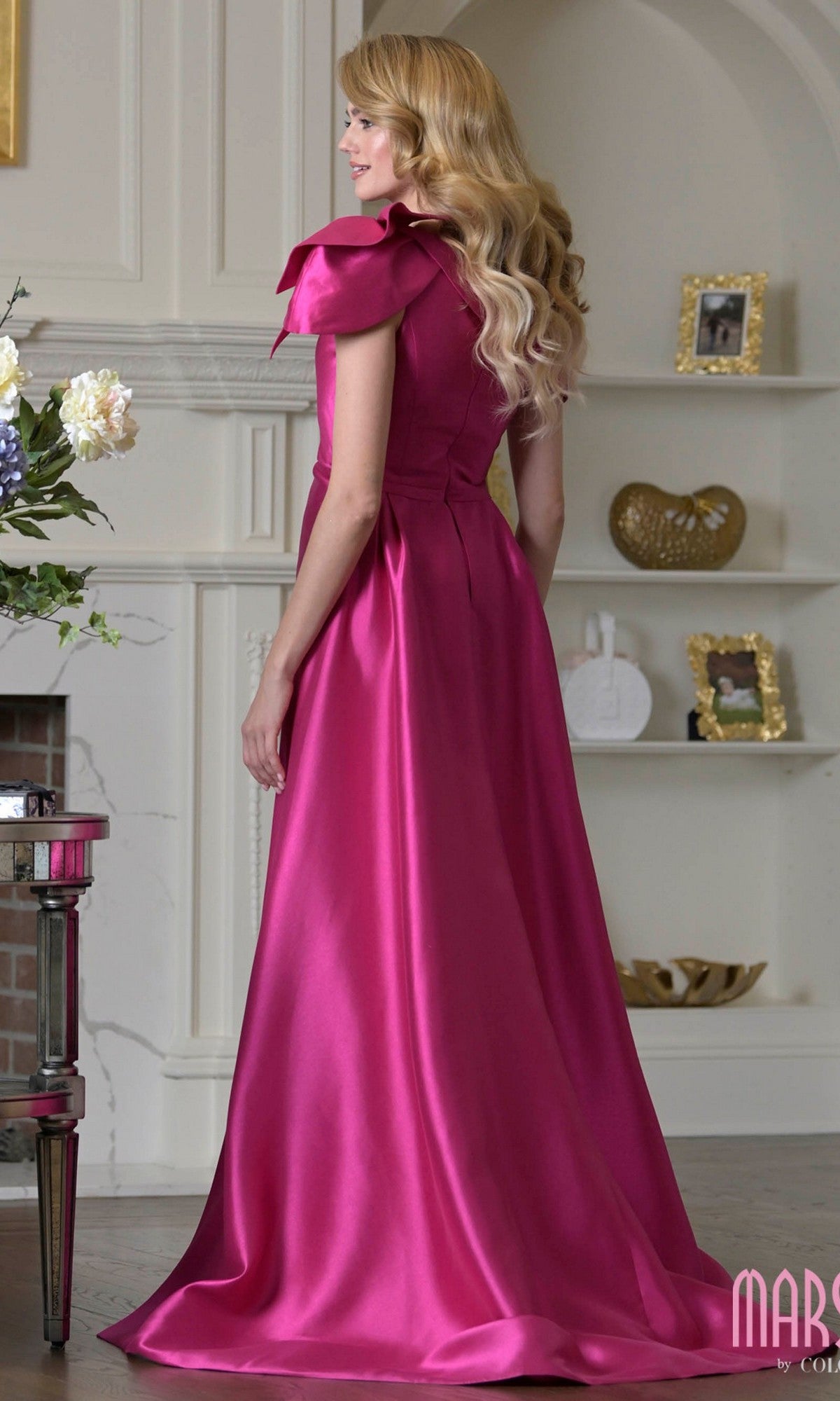 A woman with long wavy hair wears the Marsoni MV1408 long formal dress in shiny fuchsia taffeta, standing indoors and facing away from the camera beside white shelves with decorative items.