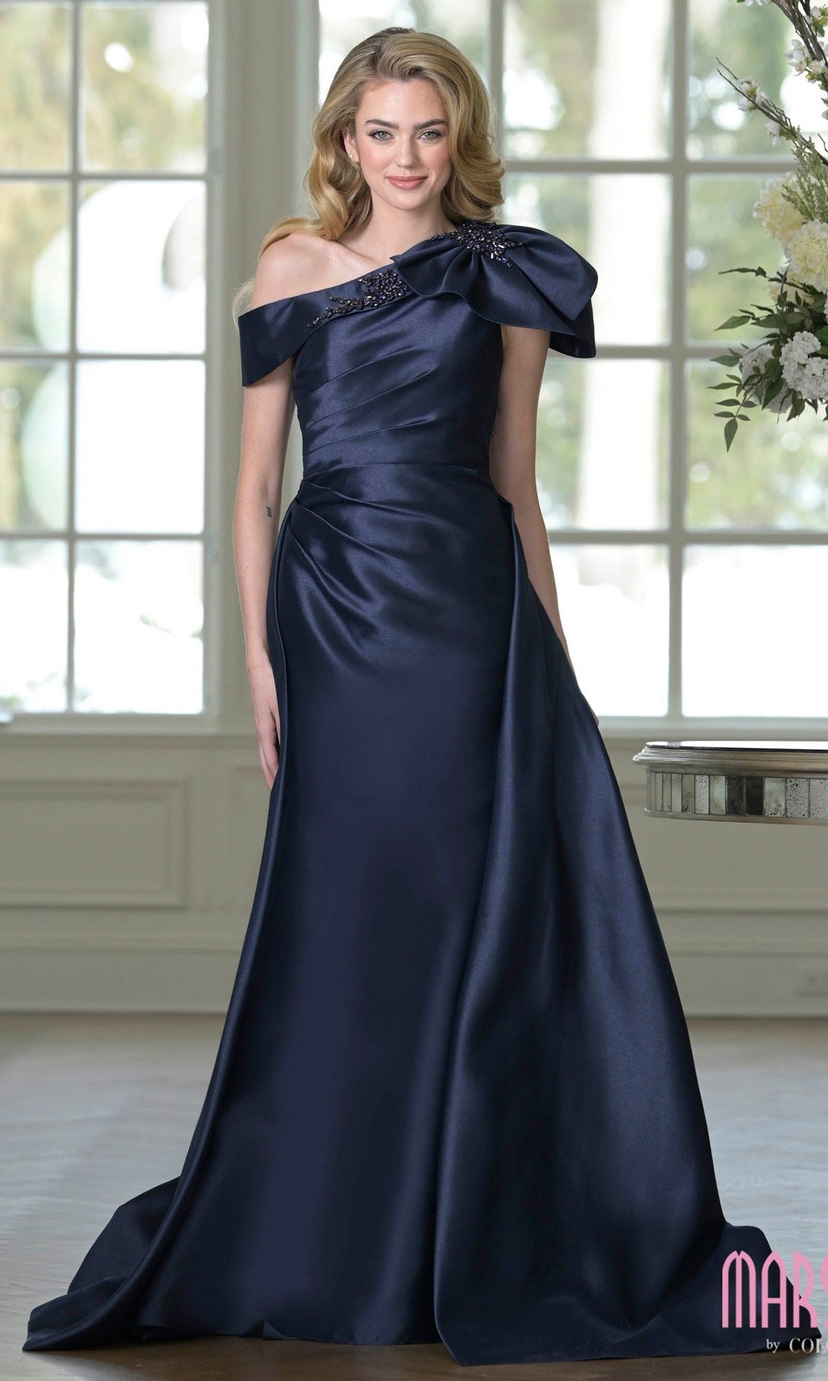 A woman stands indoors near a window and flowers, wearing the Marsoni MV1408 Long Formal Dress in Midnight, featuring a floor-length taffeta design with an asymmetrical off-the-shoulder neckline and ruched detailing.