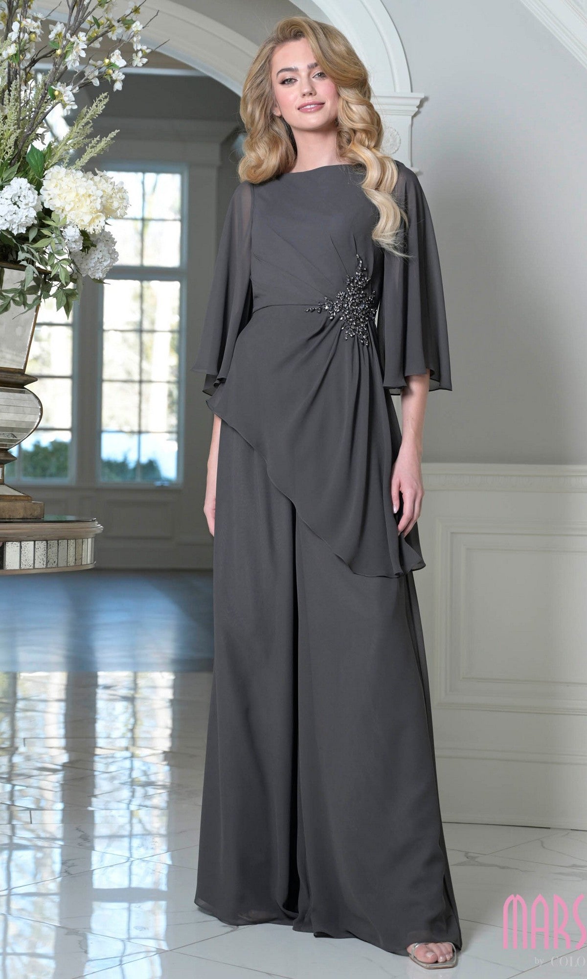 A woman stands indoors near a floral arrangement wearing the Marsoni MV1411 Mother-of-the-Bride pantsuit in charcoal, featuring wide-leg chiffon pants and draped layers.