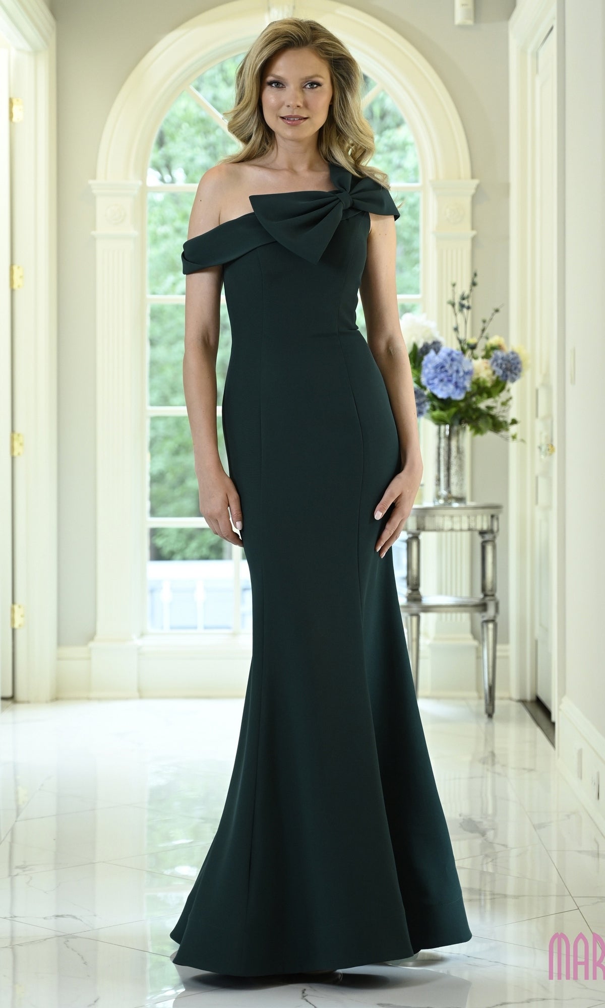 Woman wearing a floor-length, deep green Marsoni MV1432 evening gown with an asymmetrical off-the-shoulder neckline featuring a large bow detail.