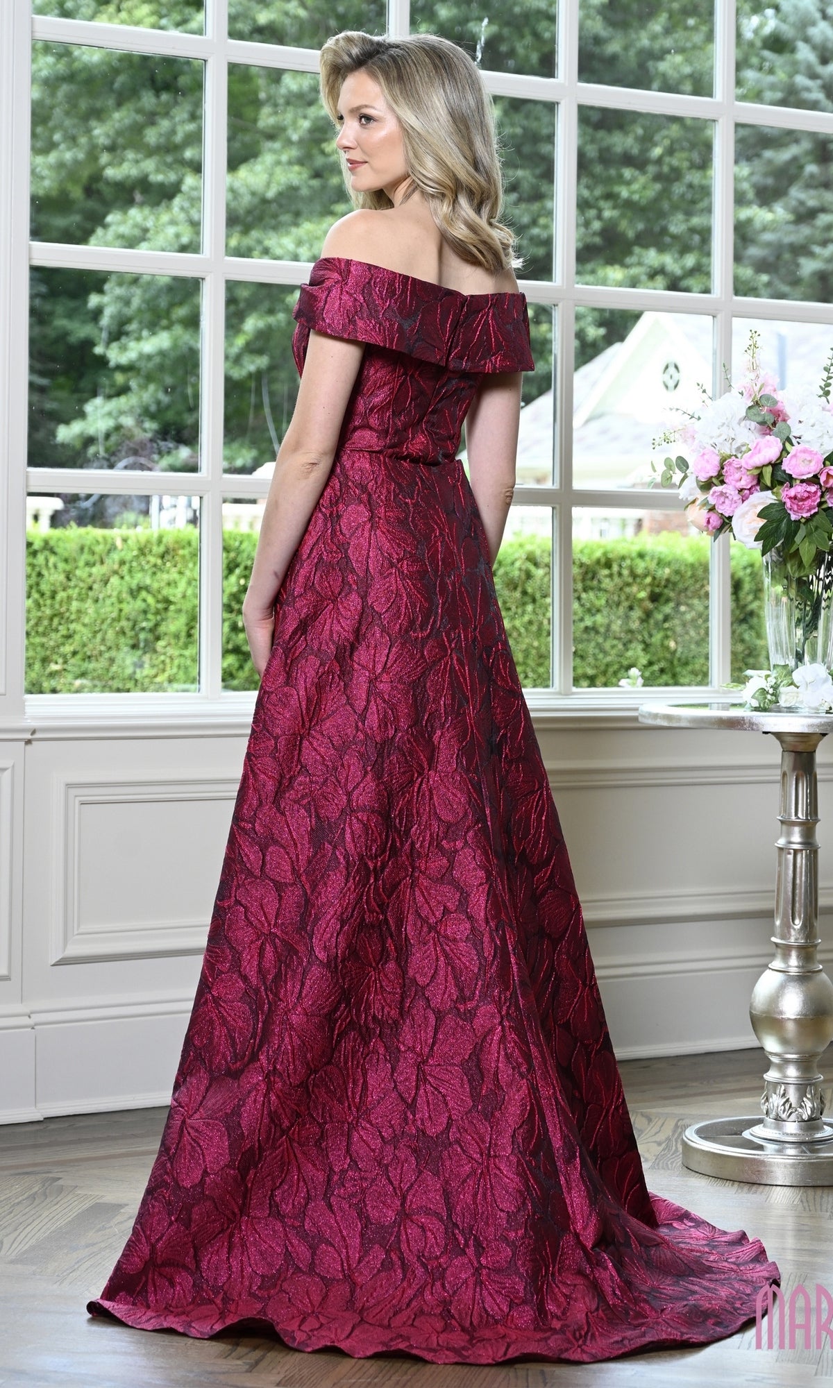 A woman wearing a Marsoni MV1440 long formal dress, an off-the-shoulder evening gown with a floor-length dark red floral jacquard pattern.