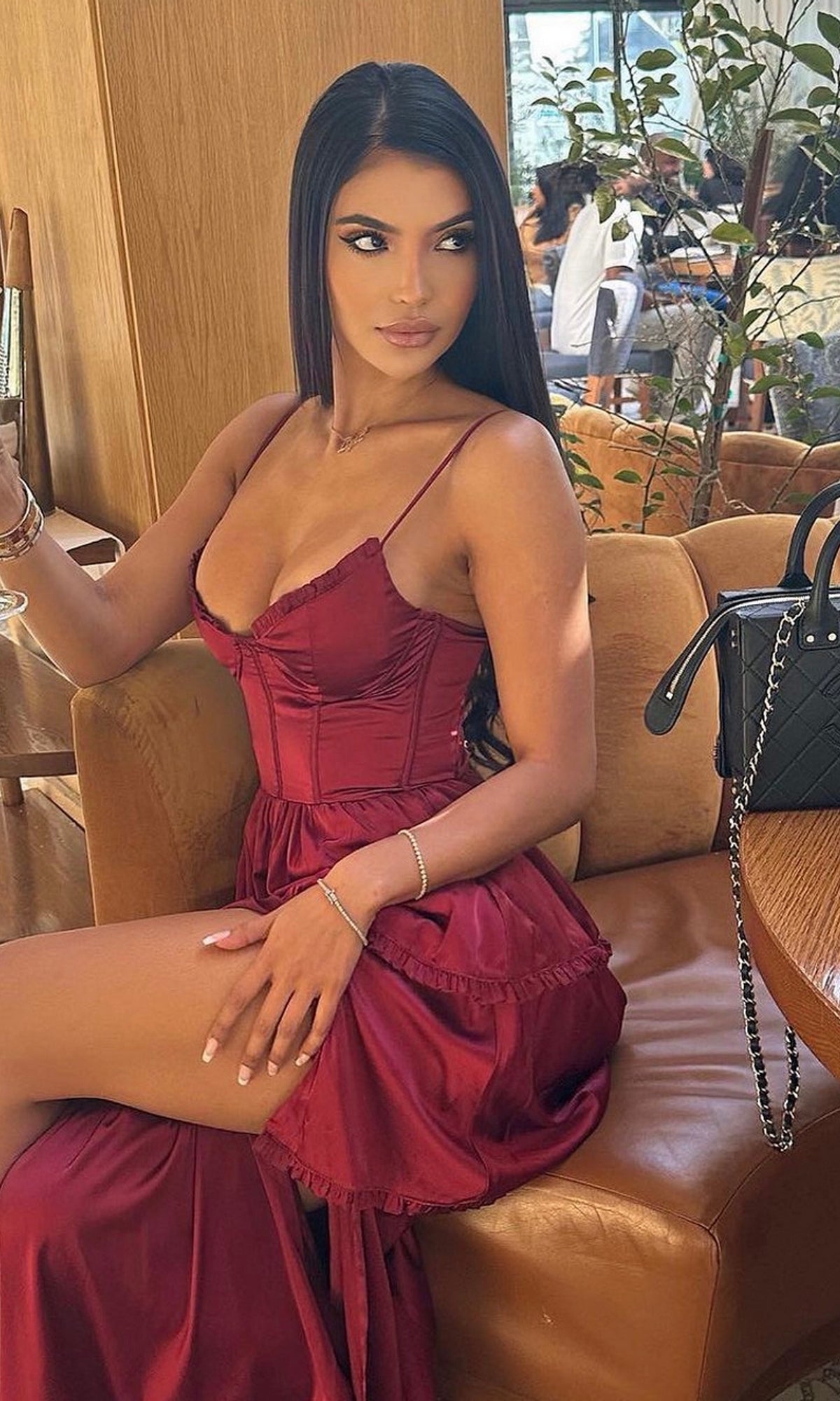 A woman with long dark hair sits on a tan chair, wearing the Velvi Lace-Up Layered Midi Party Dress Maeve in deep red. A black quilted handbag rests beside her on the seat.