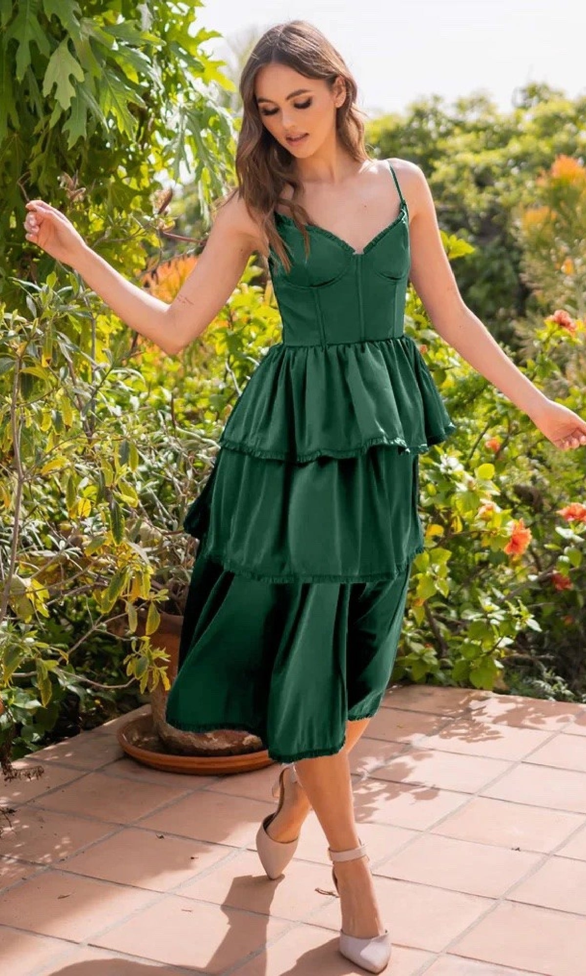 A woman poses outdoors on a tiled patio surrounded by lush greenery, wearing the Velvi Lace-Up Layered Midi Party Dress Maeve in dark green.