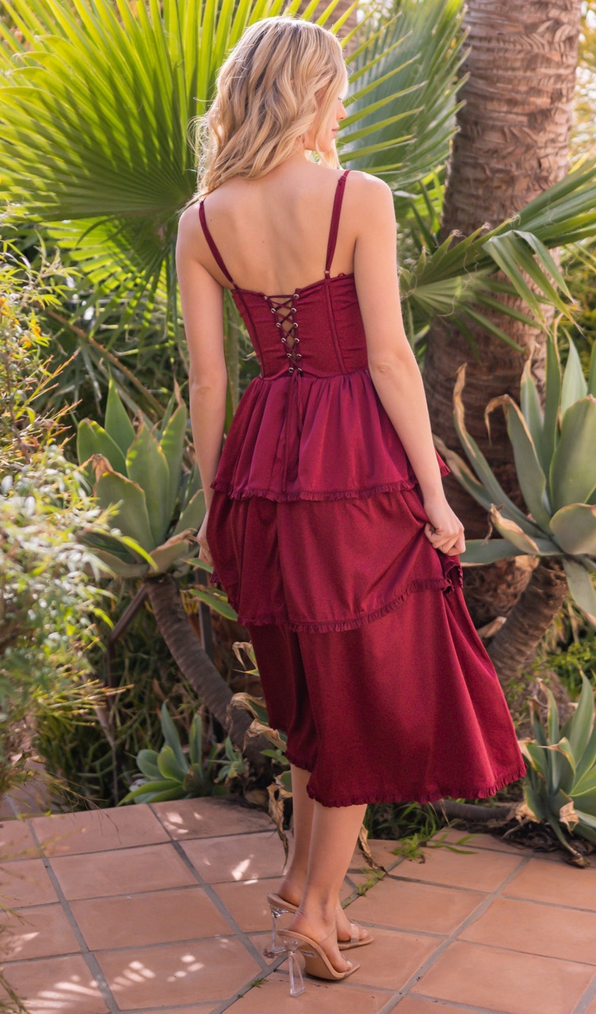 A blonde woman in the Velvi Lace-Up Layered Midi Party Dress Maeve, featuring a red tiered design and lace-up back, stands on a tiled patio amid lush tropical plants.