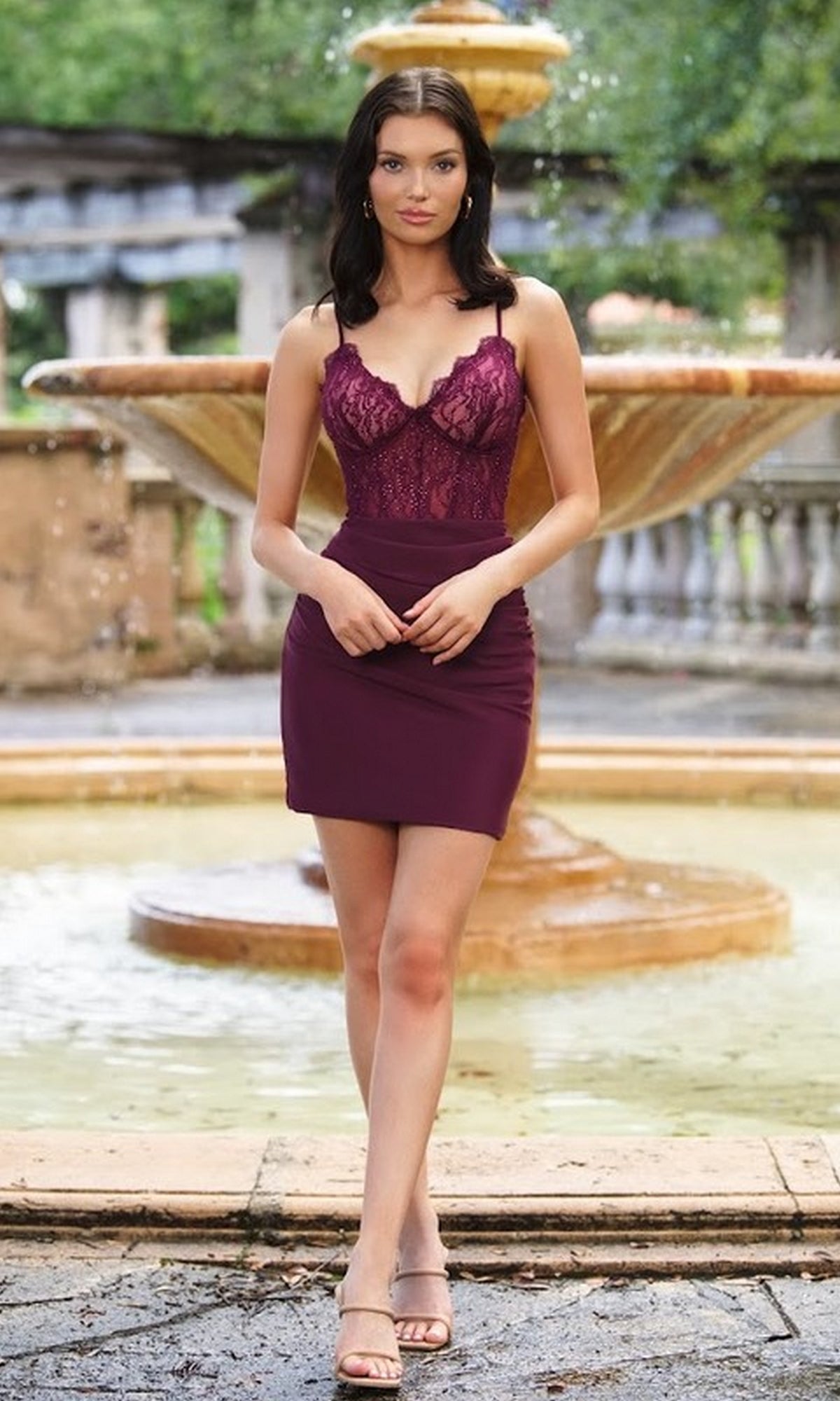A woman in a Misha Lace-Bodice Short Homecoming Dress stands outdoors in front of a stone fountain, looking at the camera.