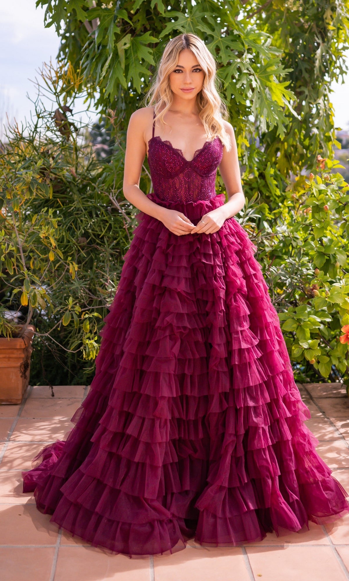 A woman stands outdoors in the Velvi Lace-Corset Long Prom Ball Gown Morgan, a deep magenta dress with ruffles, a fitted bodice, and spaghetti straps, surrounded by lush green plants.