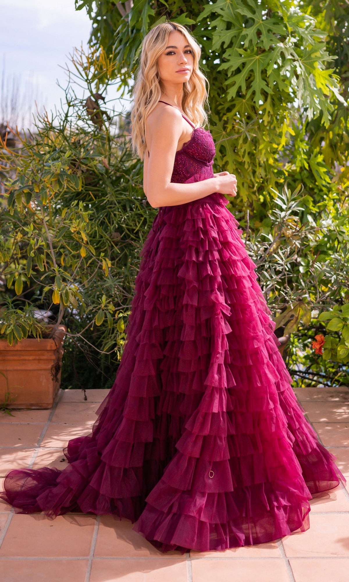 A woman stands outdoors in the Velvi Lace-Corset Long Prom Ball Gown Morgan, featuring a strapless magenta design with a fitted bodice and tiered skirt. Green foliage and tiled flooring add to the elegant atmosphere.