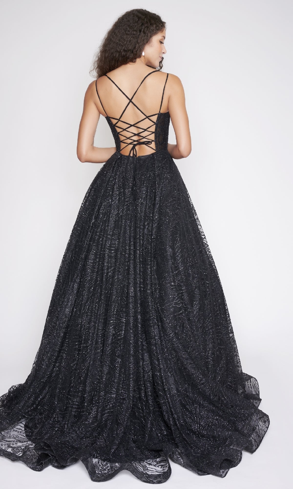 A woman models the Nina Canacci 8220 long black glitter tulle prom dress with a lace-up open back and thin crisscross straps, standing against a plain white background.