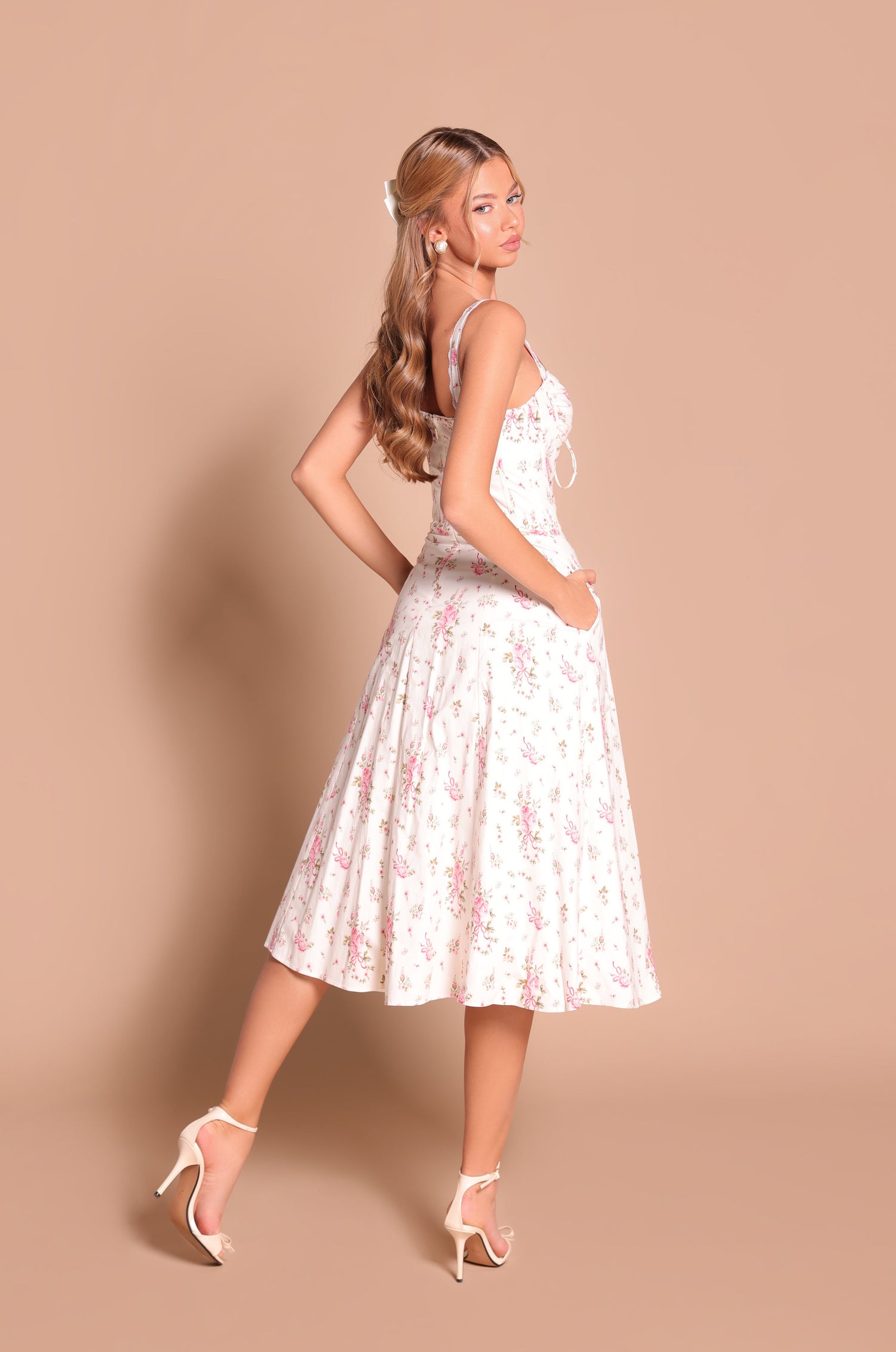 A woman wearing the Celenia Ivory White Floral Casual Midi Dress and high heels stands sideways on a beige background, looking over her shoulder.