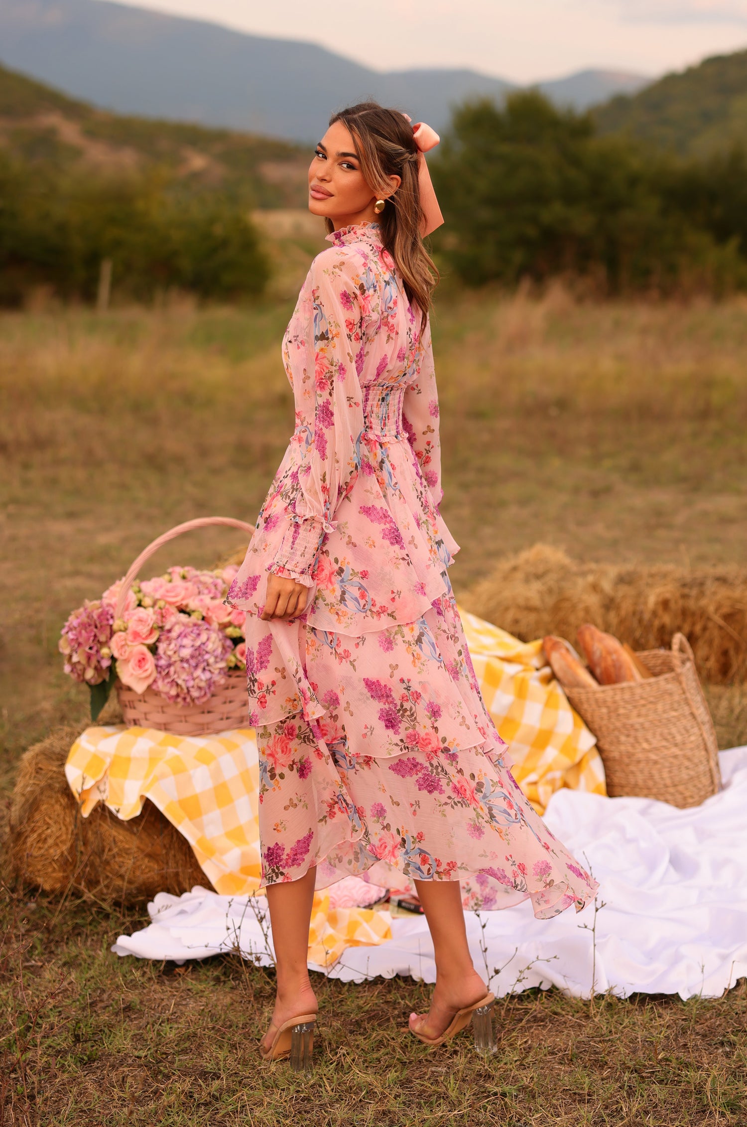 Wearing the Bellaria Long Sleeve Blush Pink Floral Maxi Dress, a woman stands on grass by a picnic setup with hay bales, flowers, and bread, surrounded by trees and mountains.