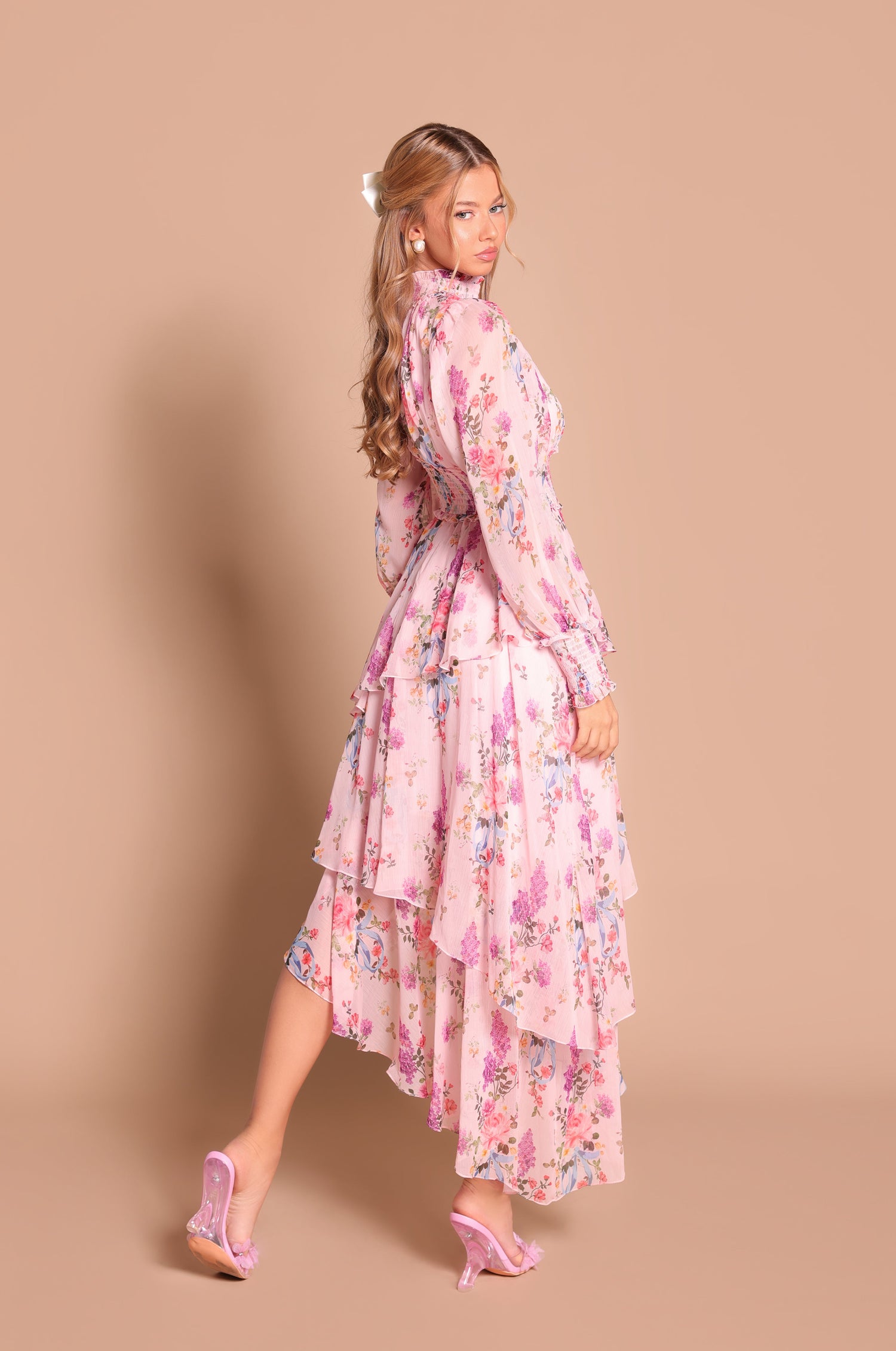 A woman in the Bellaria Long Sleeve Blush Pink Floral Maxi Dress and pink heels stands against a beige background, looking over her shoulder.