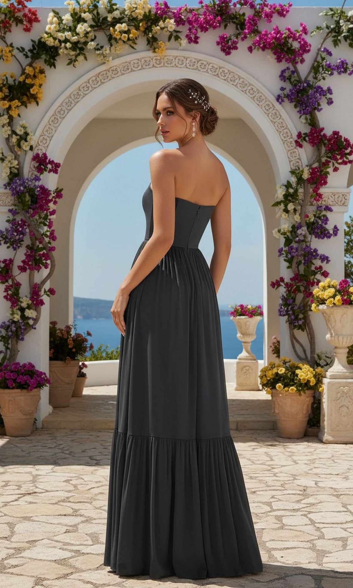 A woman in the Strapless Classic Prom Dress: PromGirl Exclusive stands on a stone patio, facing away. Surrounded by colorful flowers and potted plants, she wears the floor-length dark gray gown with a graceful a-line silhouette.