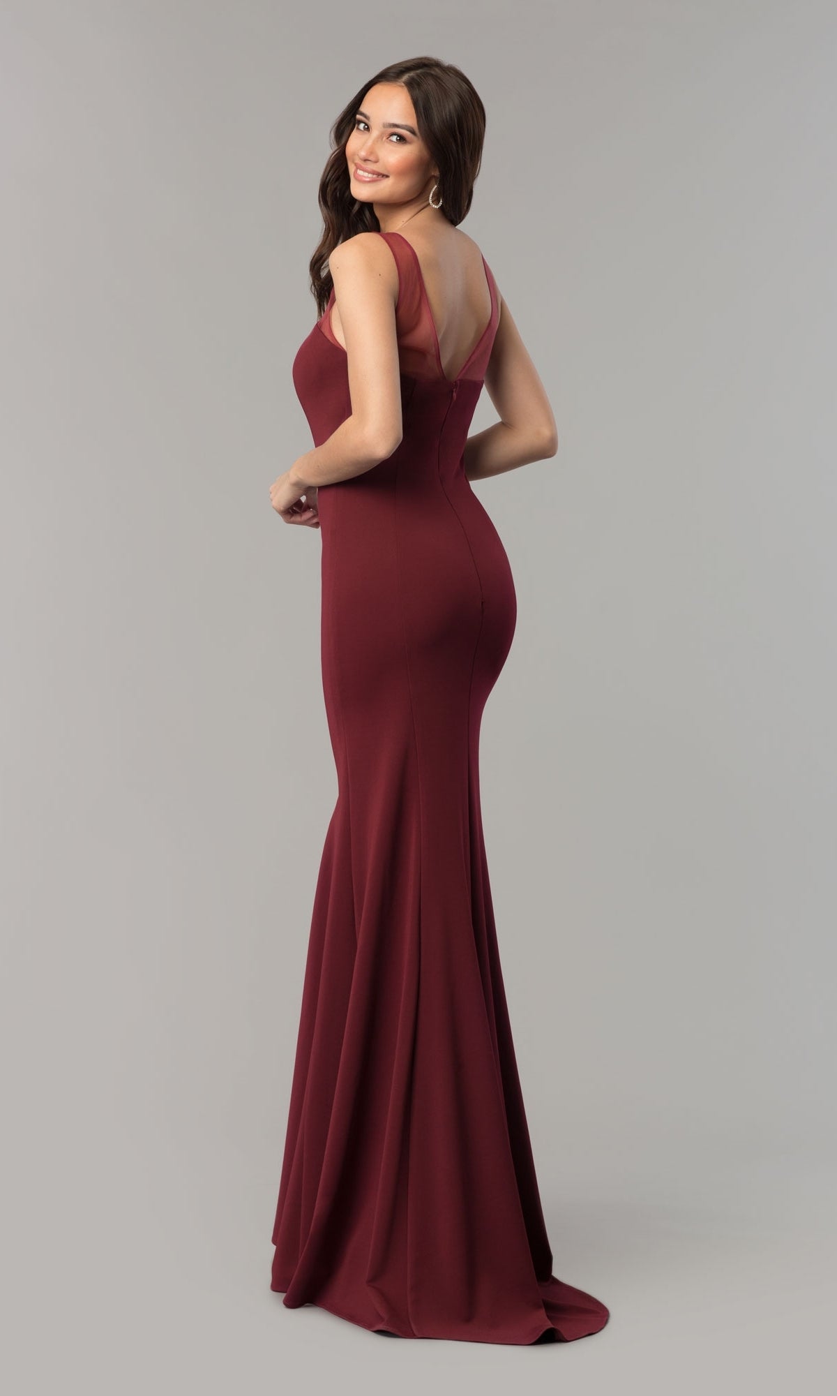A woman in the Illusion-Neckline Long Jersey Formal Prom Dress 8148—sleeveless, floor-length, and maroon with a low back—stands on a gray background, facing away and looking over her shoulder.