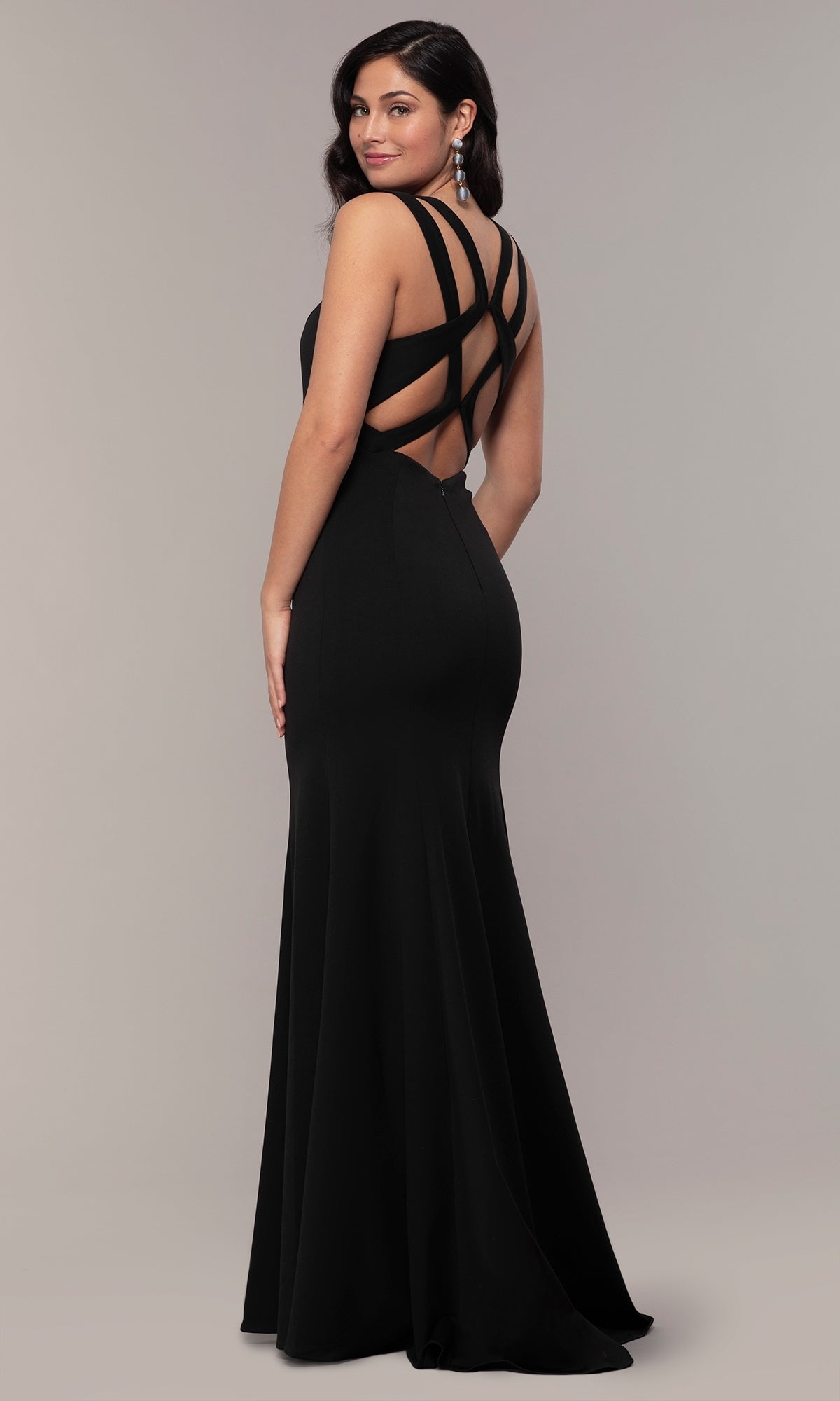 Model wearing the Strappy-Open-Back Long Formal Prom Dress 8232, featuring a fitted silhouette and elegant black design, stands against a plain background looking over her shoulder.