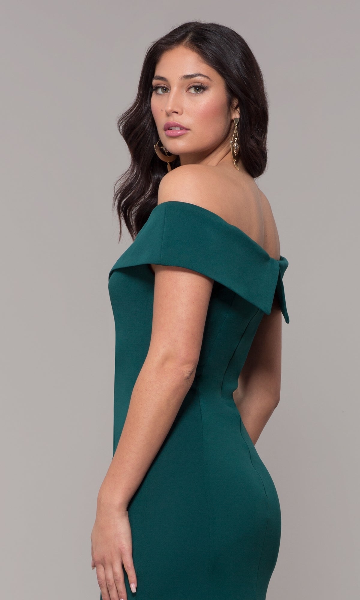 Woman in the Classic Off-the-Shoulder Formal Prom Dress 8258, a fitted emerald green jersey gown, stands against a plain gray background, glancing over her shoulder.
