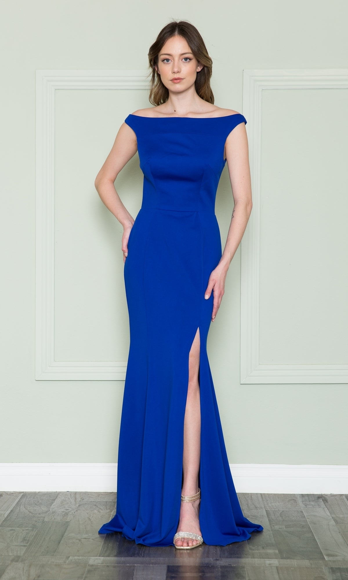 A woman poses in the Off-the-Shoulder Classic Long Prom Dress 8724 in royal, featuring a fitted silhouette and thigh-high slit, paired with gold strappy heels against a light green wall and wood floor.