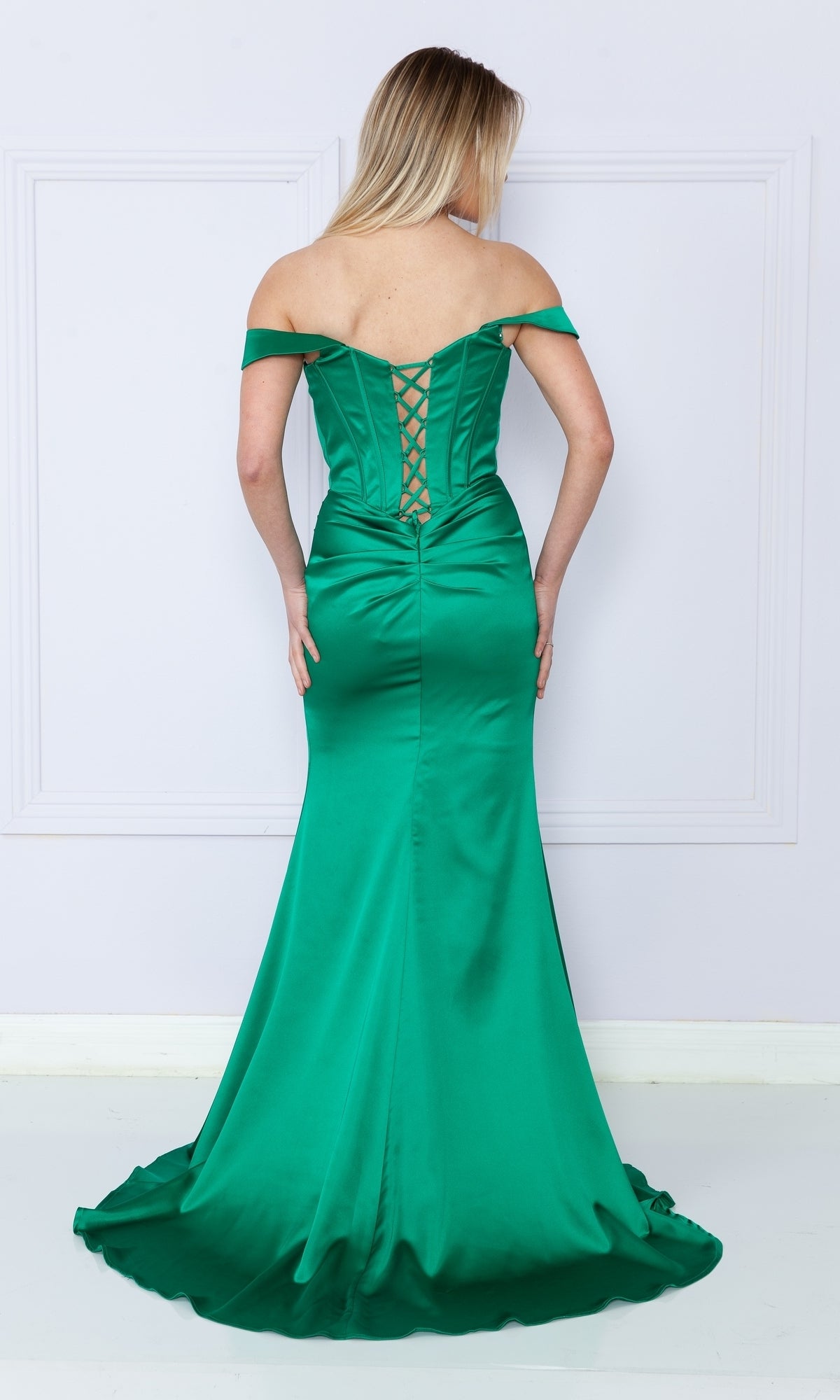 Woman in the Off-the-Shoulder Long Corset Prom Dress 9024, featuring a lace-up back, stands against a light wall, facing away from the camera.