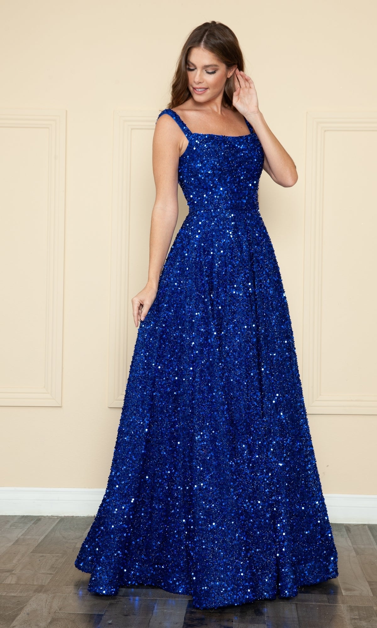 A woman stands indoors wearing the Velvet-Sequin Long A-Line Prom Gown (Poly USA 9106), a sleeveless, floor-length blue dress with sequins and a square neckline, looking down and touching her hair.