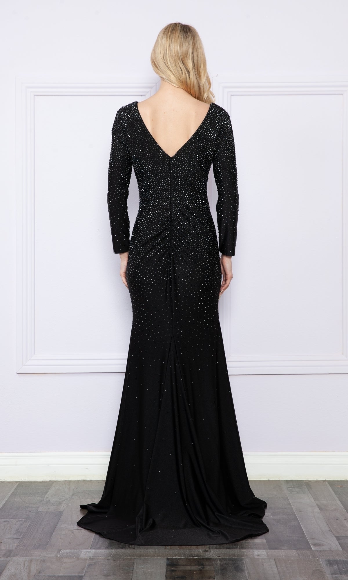 A woman stands facing away, wearing the Poly USA 9114 Long Sleeve Long Beaded Prom Dress with a deep V-back and subtle embellishments, set against a light-colored wall.