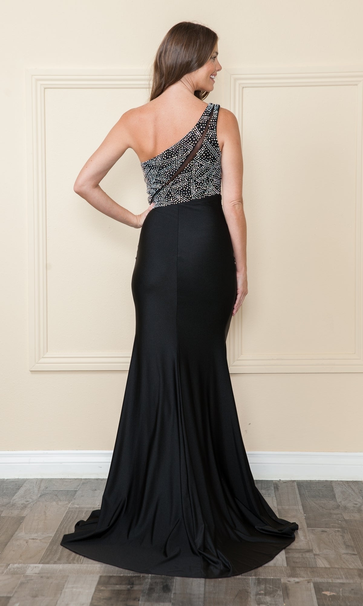 A woman stands with her back to the camera in front of a beige wall, wearing the floor-length Poly USA 9116 one-shoulder long jersey prom dress featuring a beaded top.