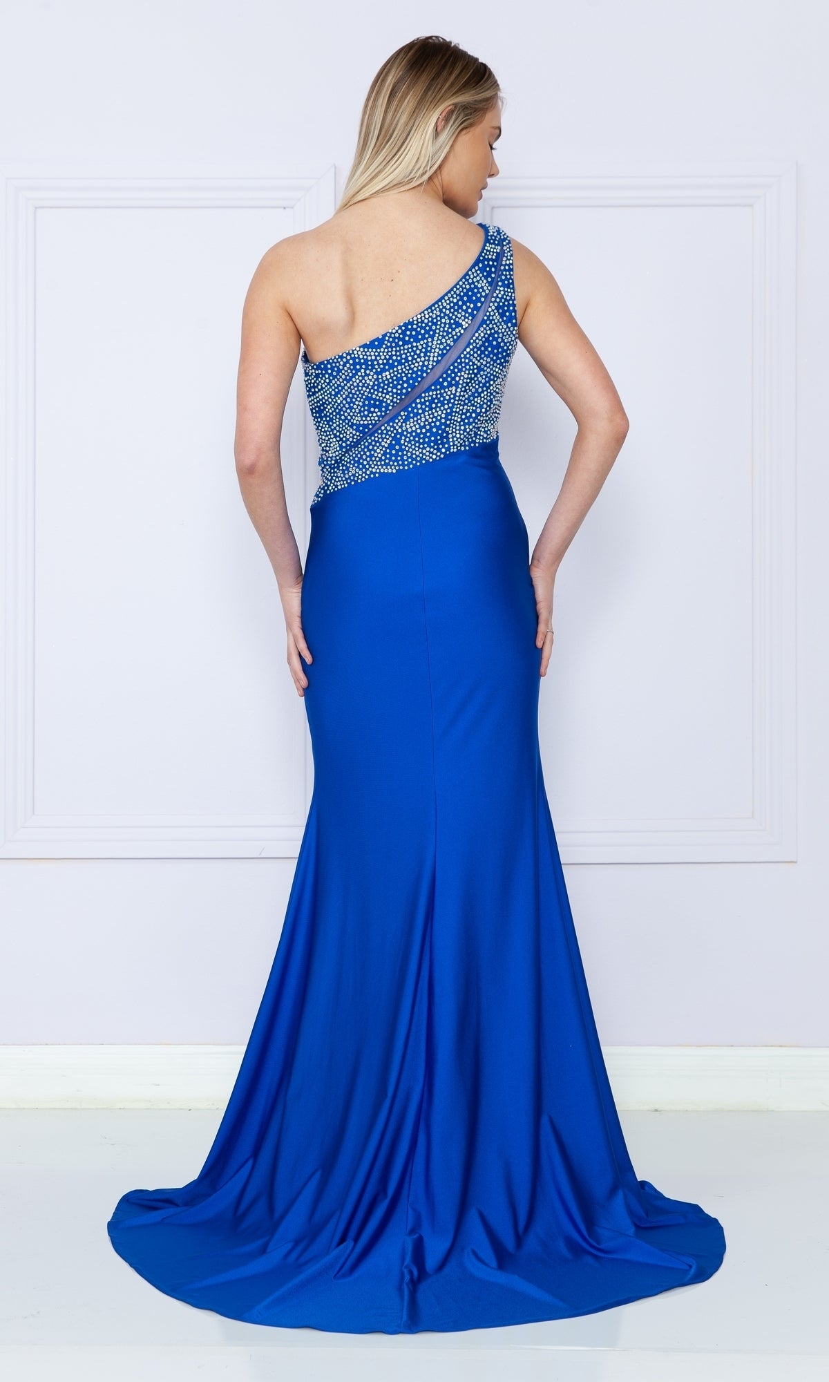 A woman wearing the Poly USA 9116 One-Shoulder Long Jersey Prom Dress with a beaded bodice, in blue, stands facing away from the camera against a light-colored wall.
