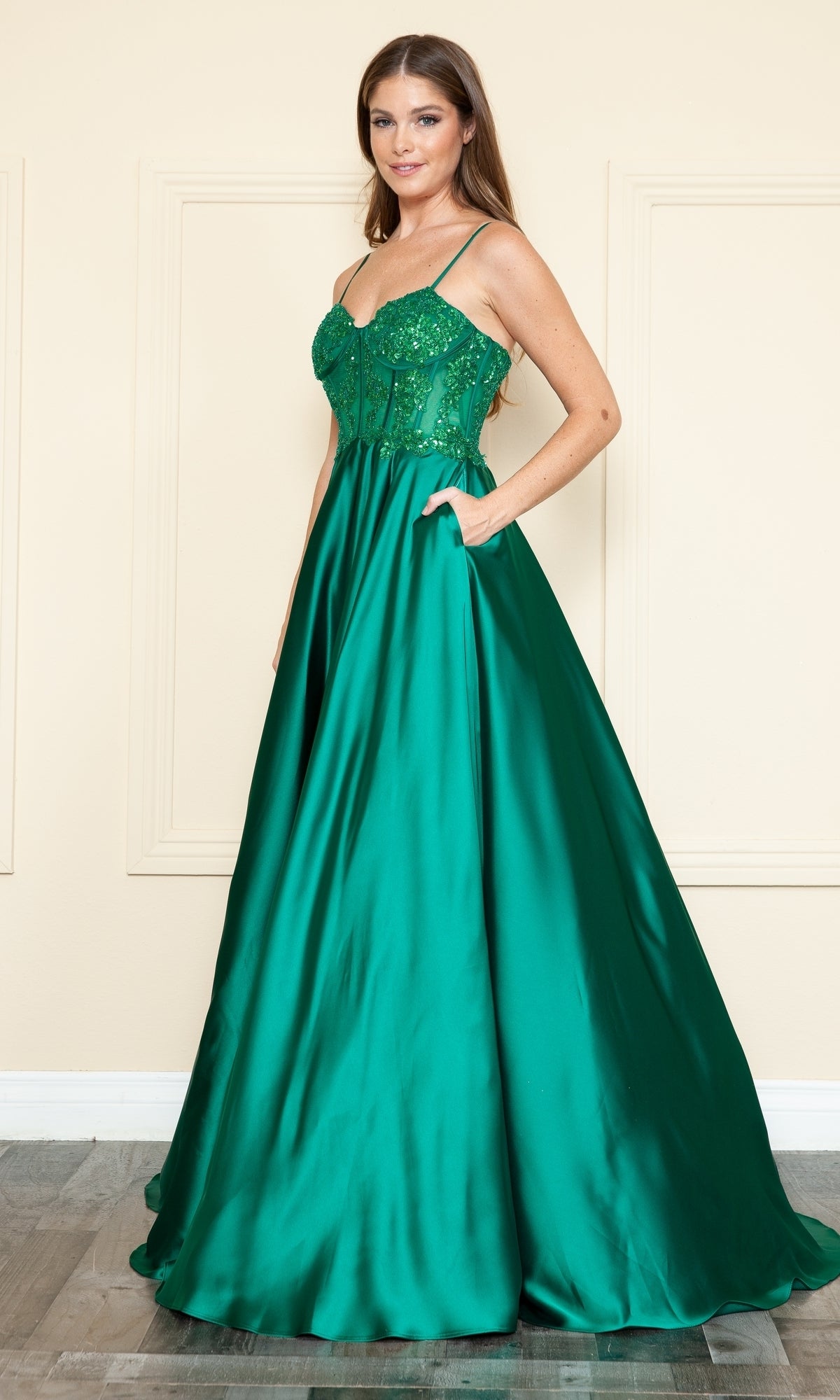 A woman stands indoors wearing the Sheer-Corset Long Satin Prom Dress (Poly USA 9126), a floor-length emerald green satin gown with a fitted, embellished bodice and thin straps.