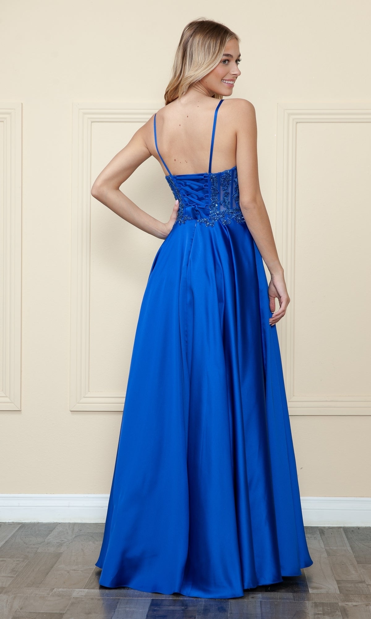 A woman stands facing away, wearing the Sheer-Corset Long Satin Prom Dress (Poly USA 9126) in royal blue, featuring thin straps and a fitted, beaded corset bodice.