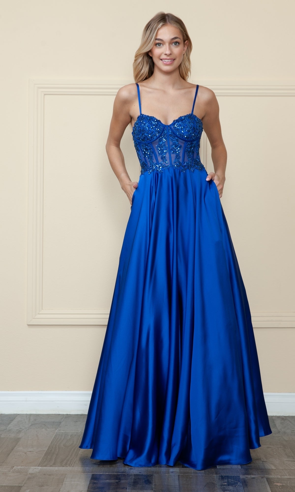 A woman wearing the Sheer-Corset Long Satin Prom Dress by Poly USA 9126 in royal blue, featuring thin straps, an embroidered corset bodice, and a flowing satin skirt, stands against a light-colored wall.
