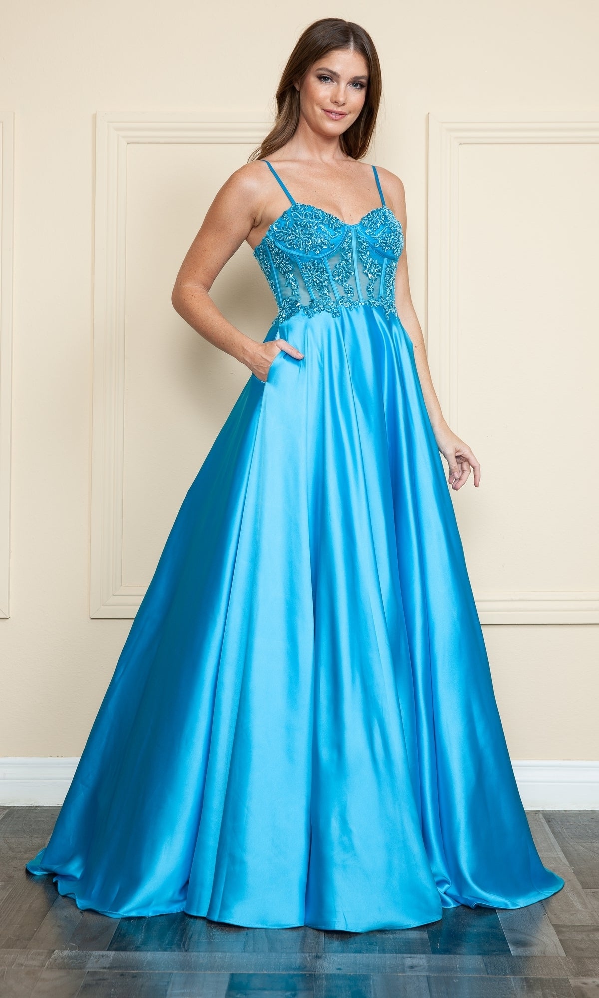 A woman wearing the Sheer-Corset Long Satin Prom Dress (Poly USA 9126) with a bright blue hue, fitted beaded bodice, spaghetti straps, and flowing A-line skirt stands indoors against a beige wall.