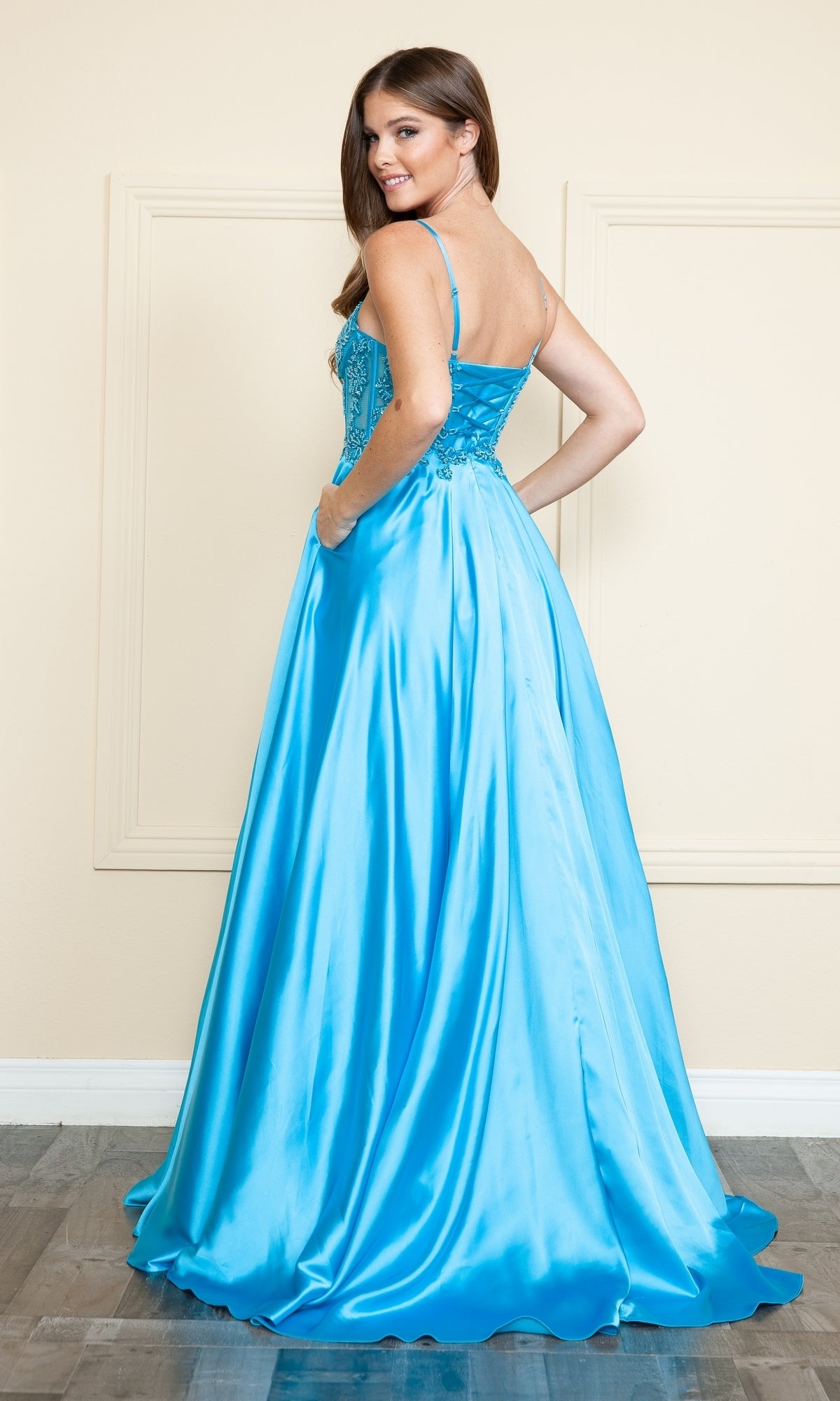 A woman wearing the Sheer-Corset Long Satin Prom Dress (Poly USA 9126) in shiny turquoise, featuring thin straps and an A-line silhouette, stands looking over her shoulder against a light-colored wall.