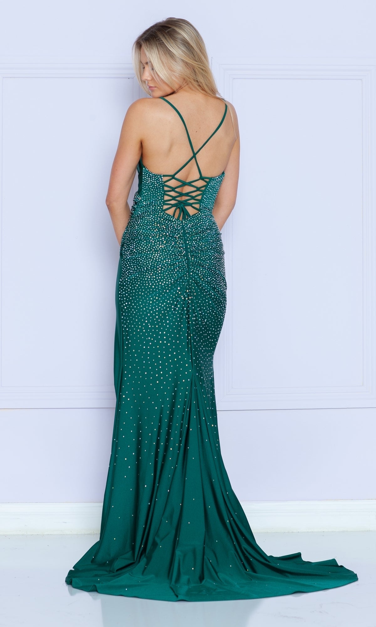 A woman stands against a light purple wall wearing the Faux-Wrap Long Beaded Prom Dress (Poly USA 9144), a floor-length, emerald green gown with sparkling embellishments and a lace-up back.
