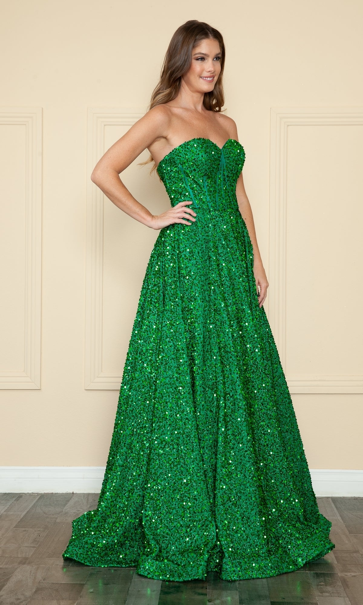 A woman stands indoors wearing the Poly USA 9152 Strapless Sequin Prom Ball Gown—an emerald green, floor-length dress with a fitted bodice and full, sweeping skirt.