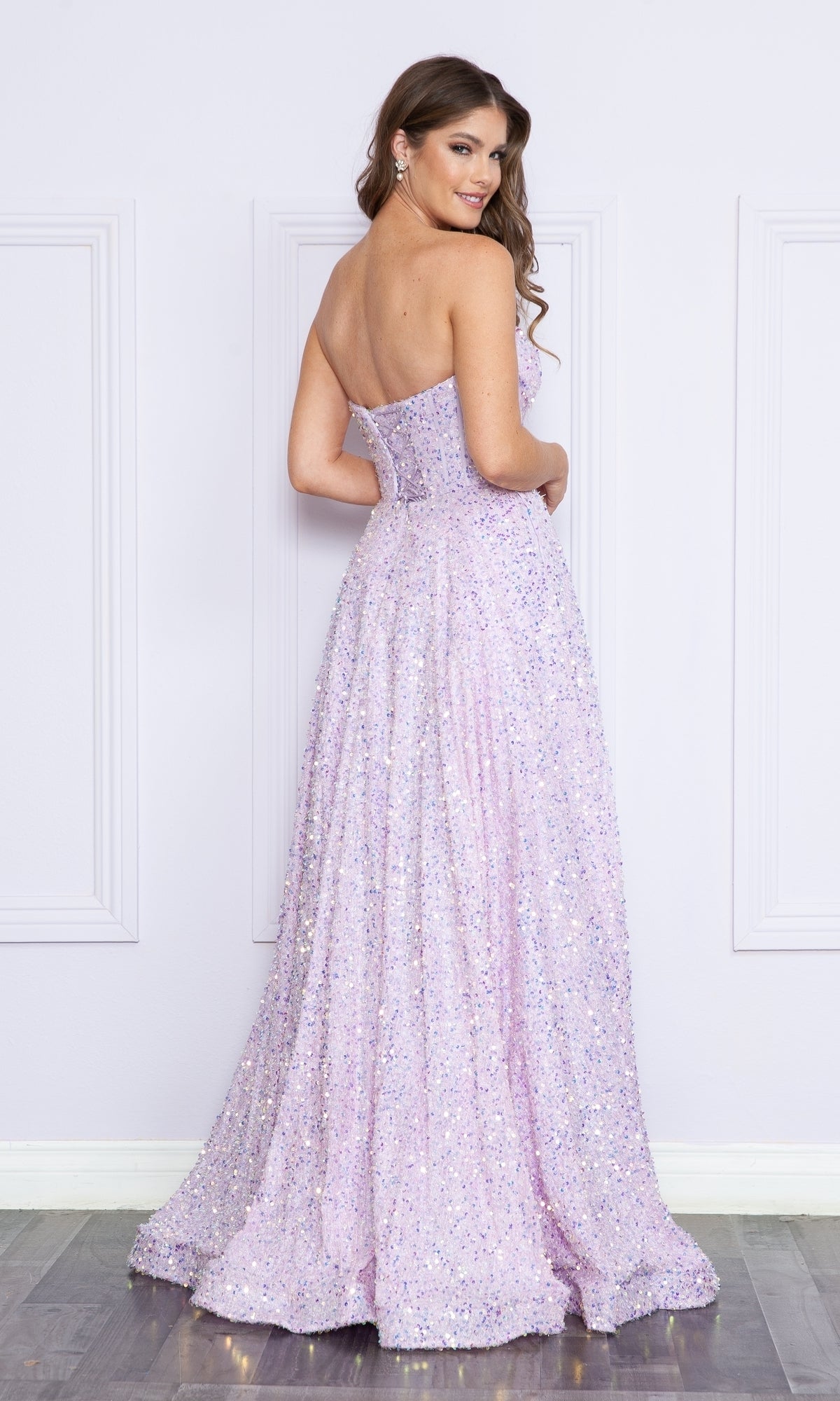 A woman in the Strapless Sequin Prom Ball Gown: Poly USA 9152 stands with her back to the camera, glancing over her shoulder against a light wall.