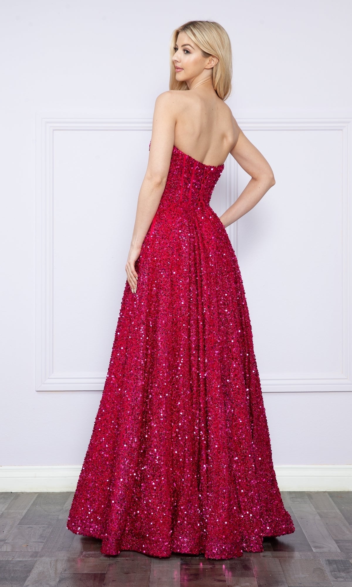 A woman stands on a wooden floor, facing away, wearing the Poly USA 9152 Strapless Sequin Prom Ball Gown—a floor-length, bright pink dress—against a light-colored wall.