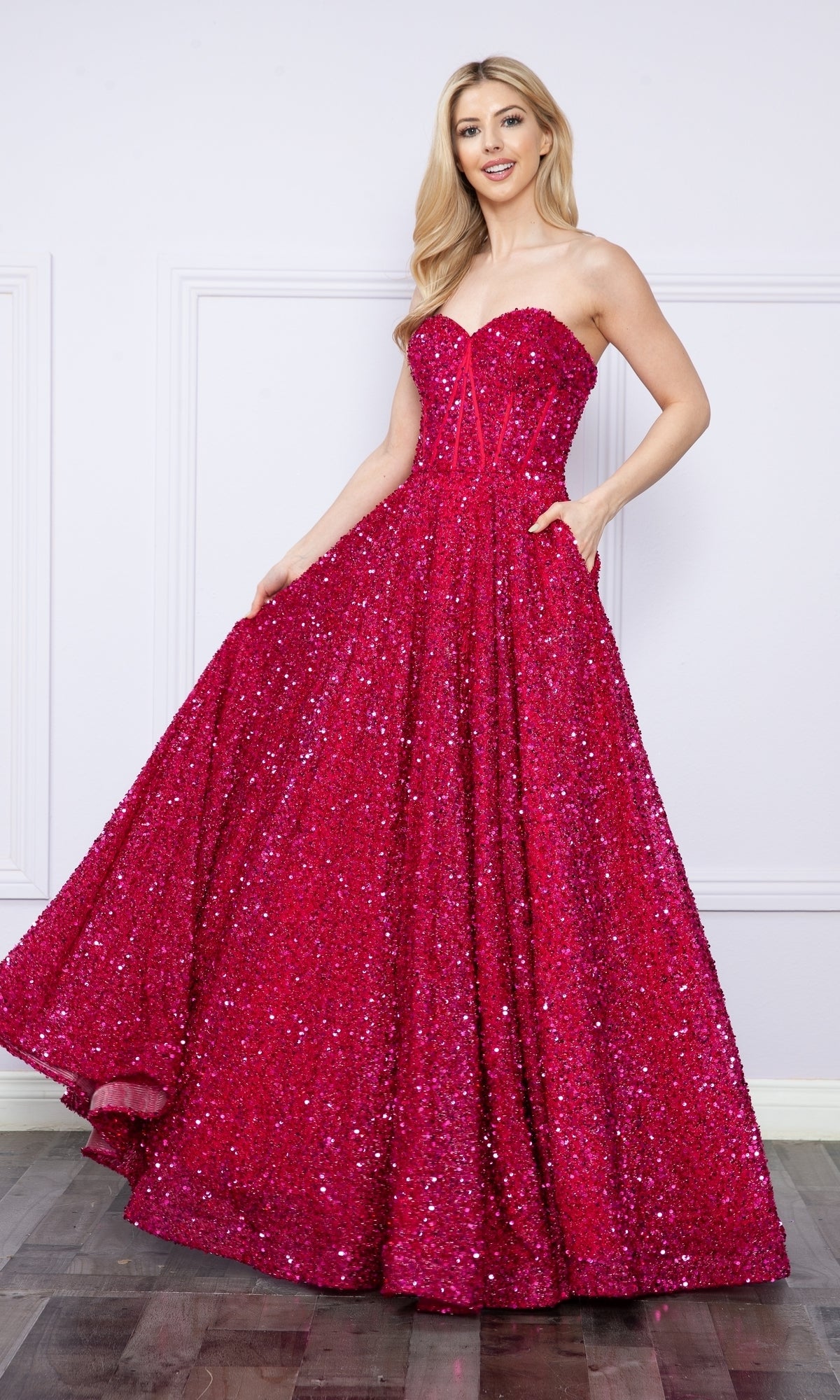 A woman stands indoors on a wooden floor against a white wall, wearing the Poly USA 9152 Strapless Sequin Prom Ball Gown in bright pink, featuring a floor-length silhouette.