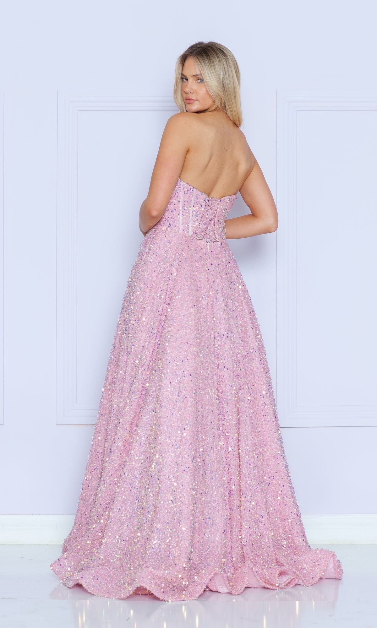A woman faces away, wearing the Strapless Sequin Prom Ball Gown: Poly USA 9152—a floor-length pink dress with a fitted bodice and flared skirt, shimmering as she stands against a light-colored wall.