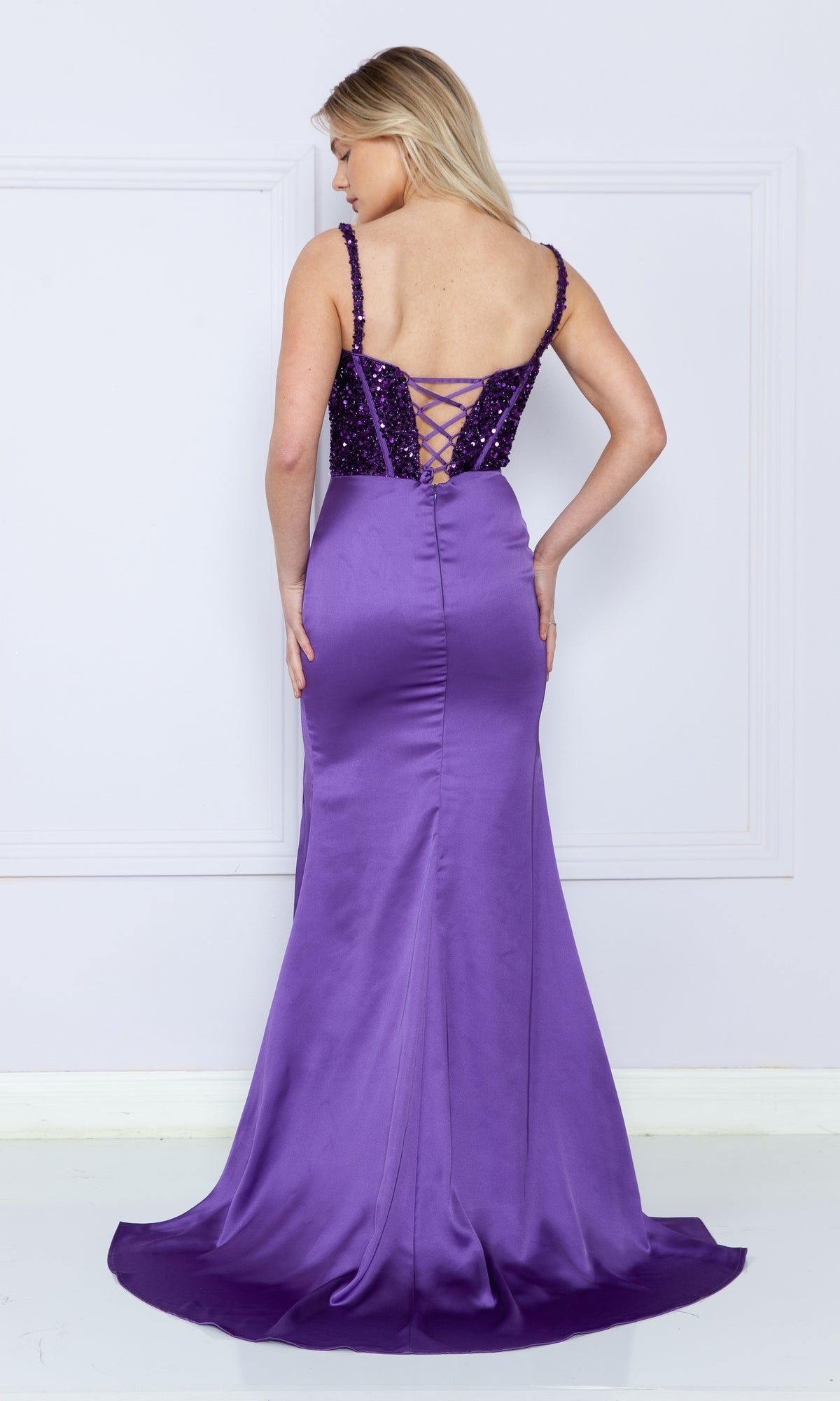 A woman in the Lace-Up Sequin-Bodice Long Satin Prom Dress 9176, featuring a corset-style sequin bodice and flowing satin skirt, stands against a light wall, facing away from the camera.
