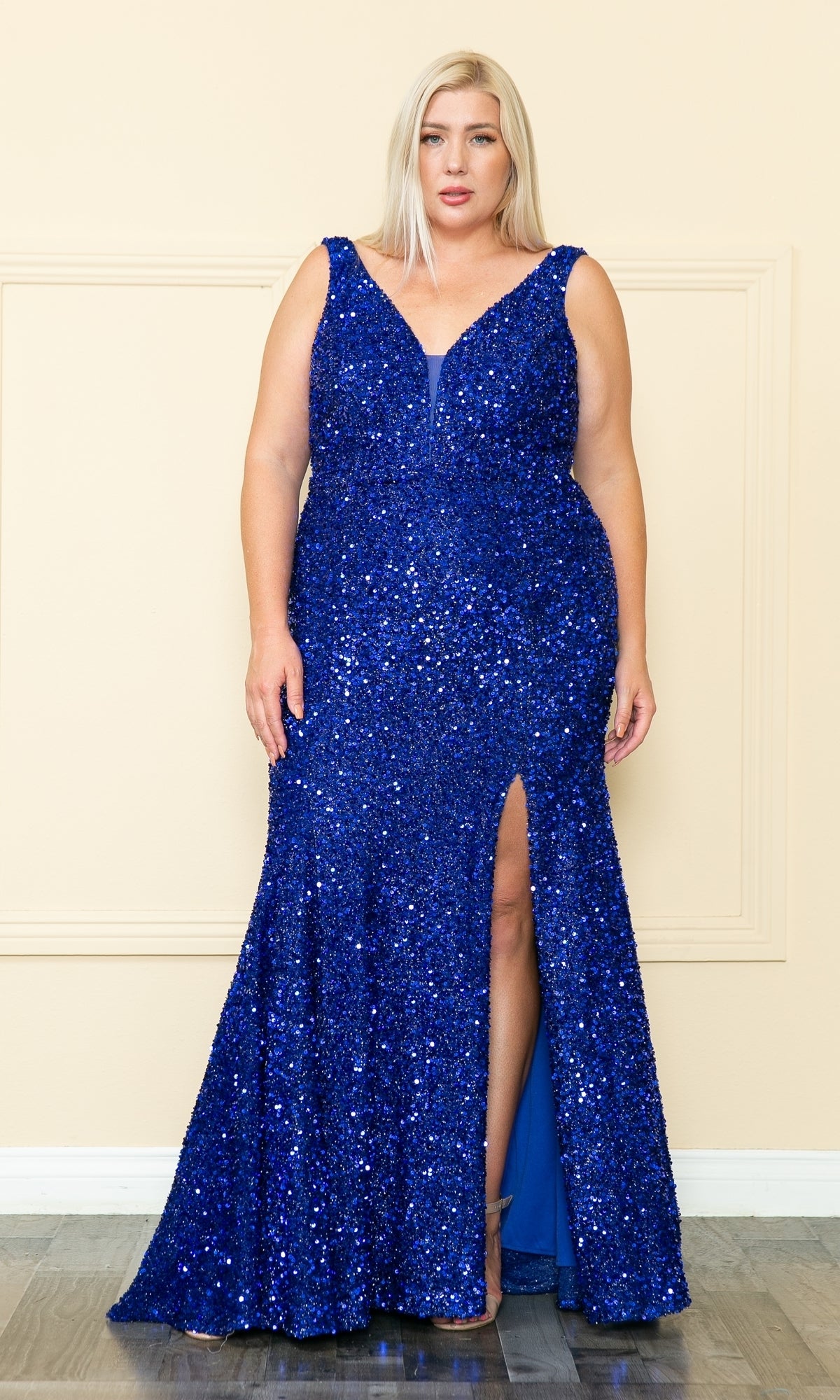 A woman stands against a beige wall wearing the royal Velvet-Sequin Plus-Size Long Prom Dress W1122, featuring a sleeveless design, deep V-neckline, and thigh-high slit.