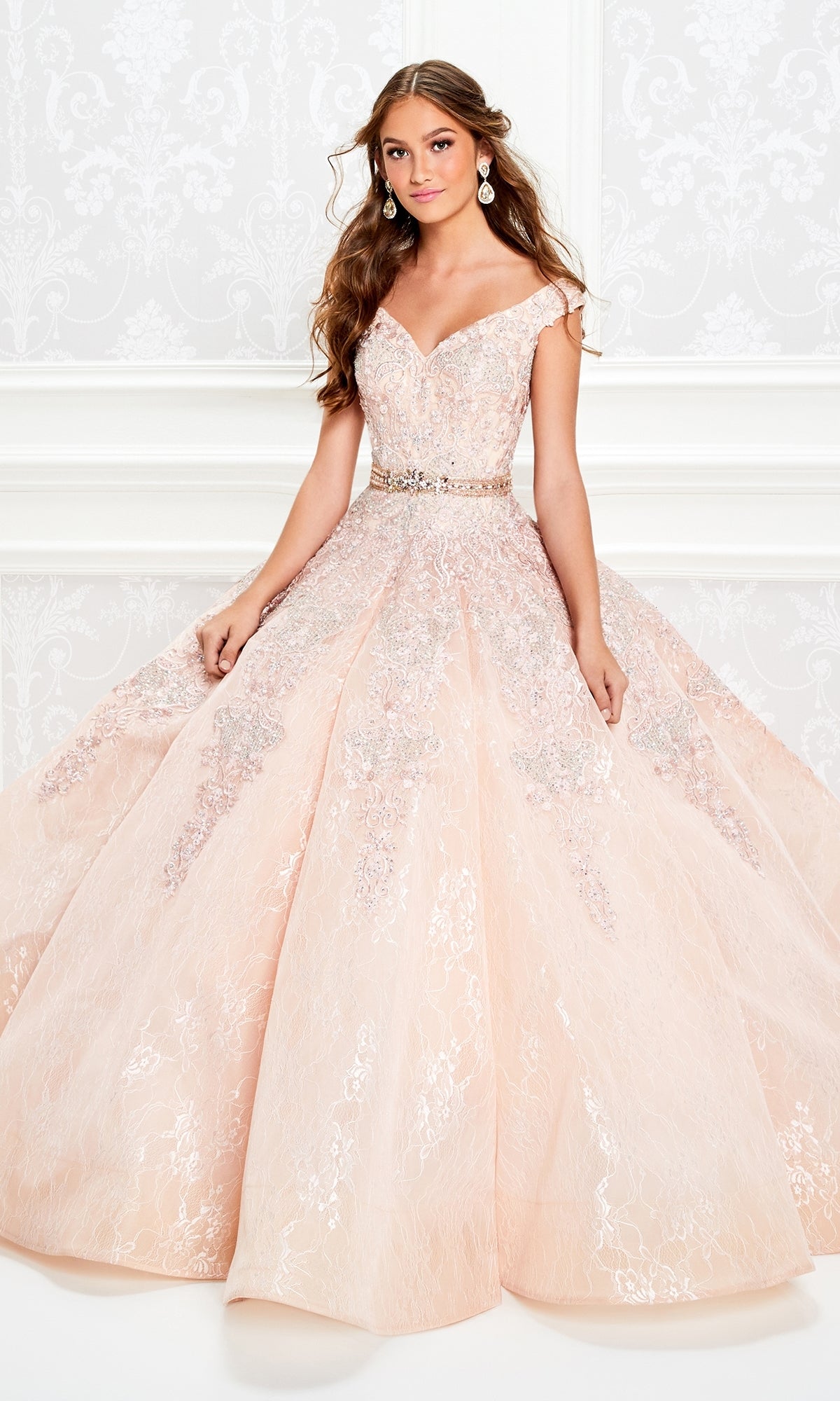 A young woman wearing the Princesa Lace Ball Gown Quinceañera Dress PR11921, an off-the-shoulder blush pink gown with silver embroidery and a beaded belt, stands in an elegant white room accessorized with drop earrings.