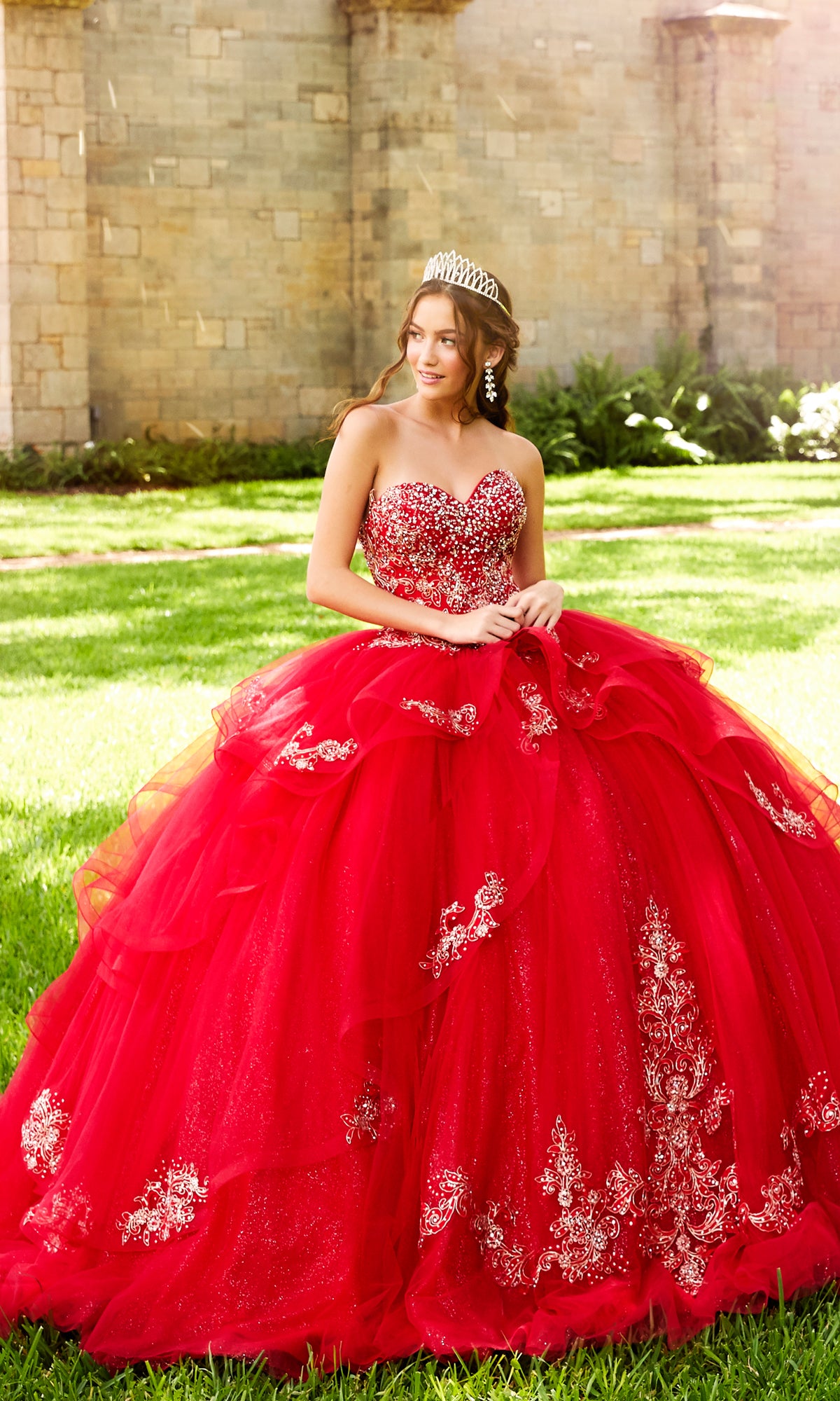 A young woman wearing the Princesa Beaded Lace Quinceañera Dress PR11923 and a tiara stands on grass before a stone wall, smiling as she looks to the side.