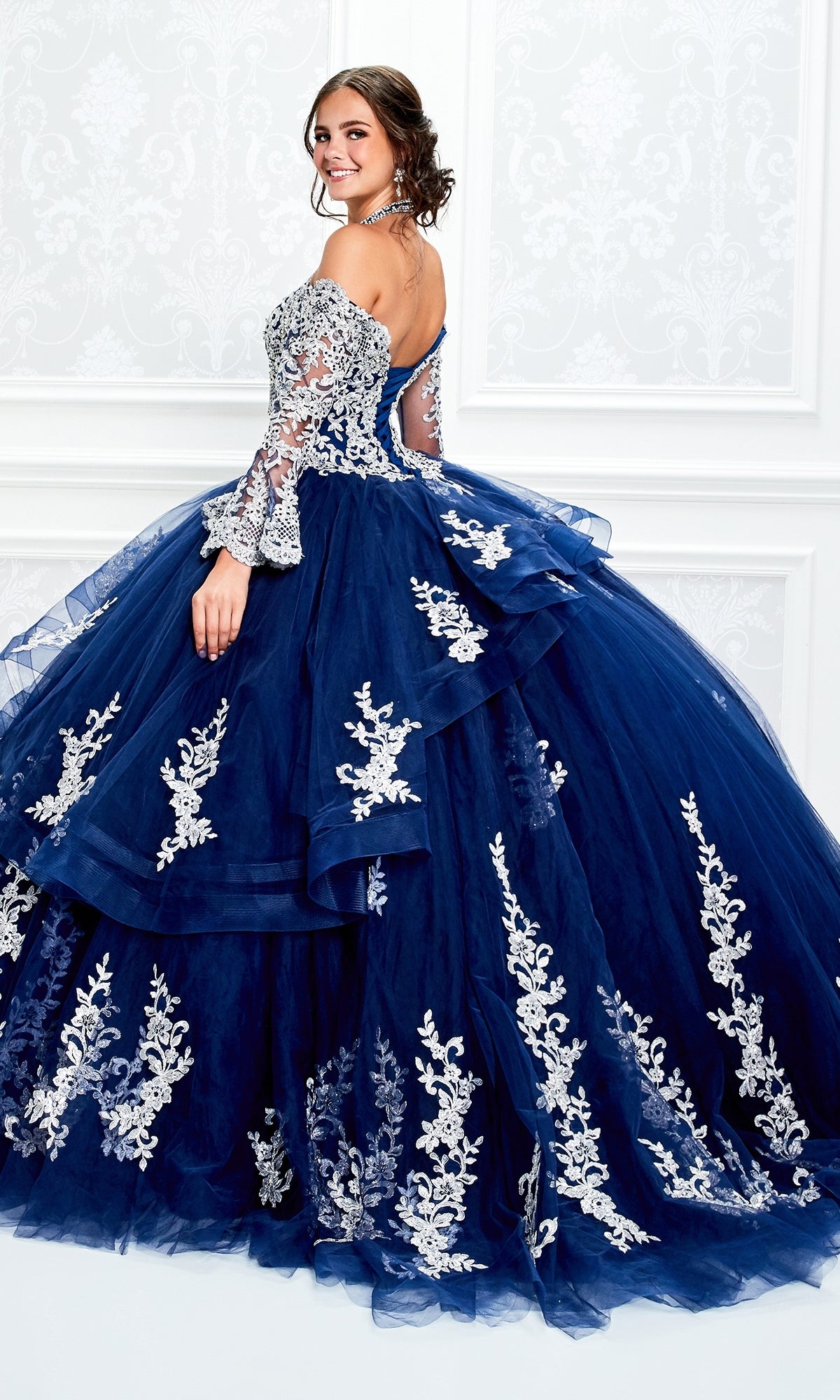 A woman poses in the Princesa Halter Ball Gown Quinceañera Dress PR11927, a blue gown with white floral embroidery, lace details, a halter neckline, and a full layered skirt.