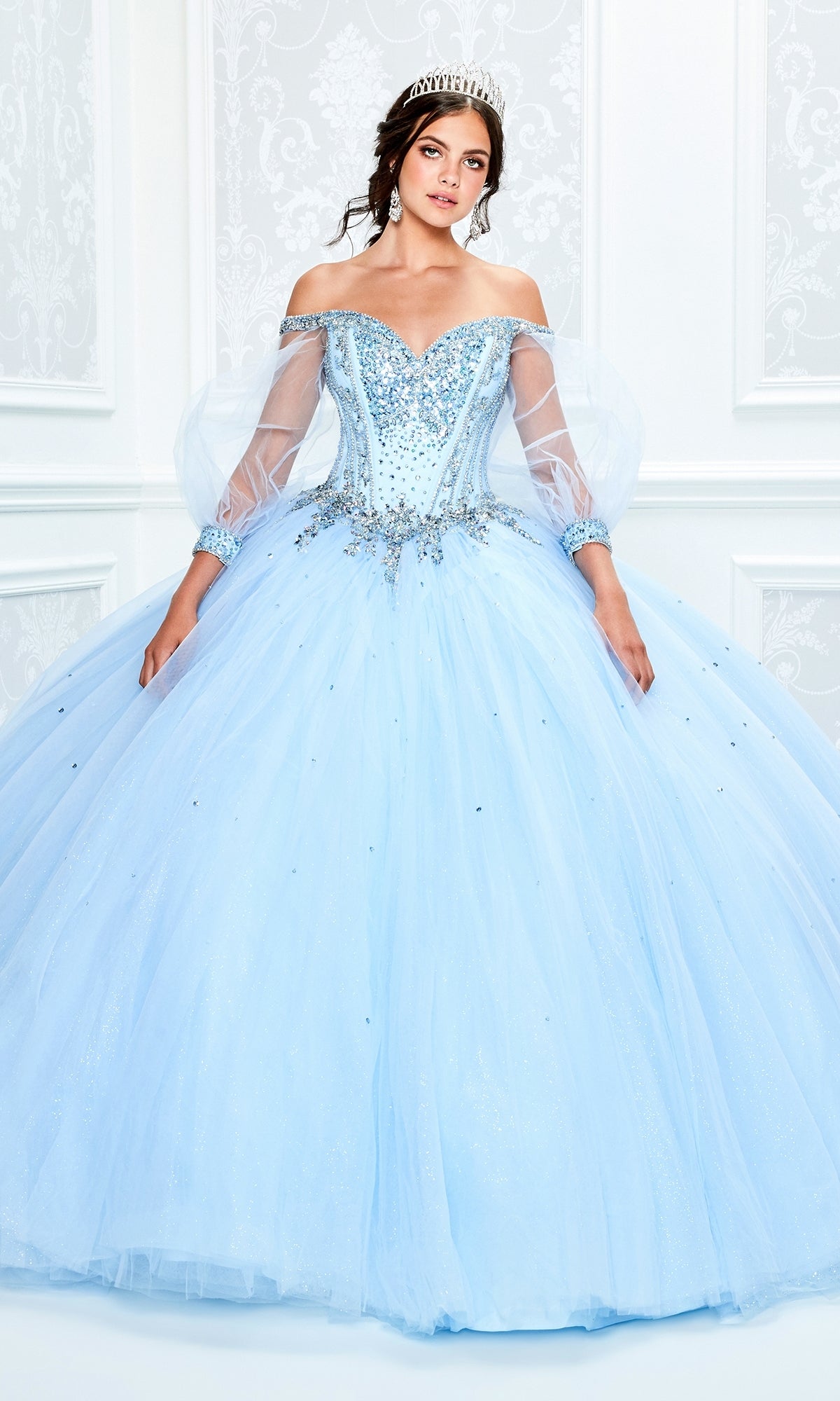 A woman wears the Princesa Light Blue Quinceañera Dress PR11937, featuring an off-the-shoulder jeweled corset bodice, full tulle skirt, sheer sleeves, and a tiara, posed against a white ornate background.