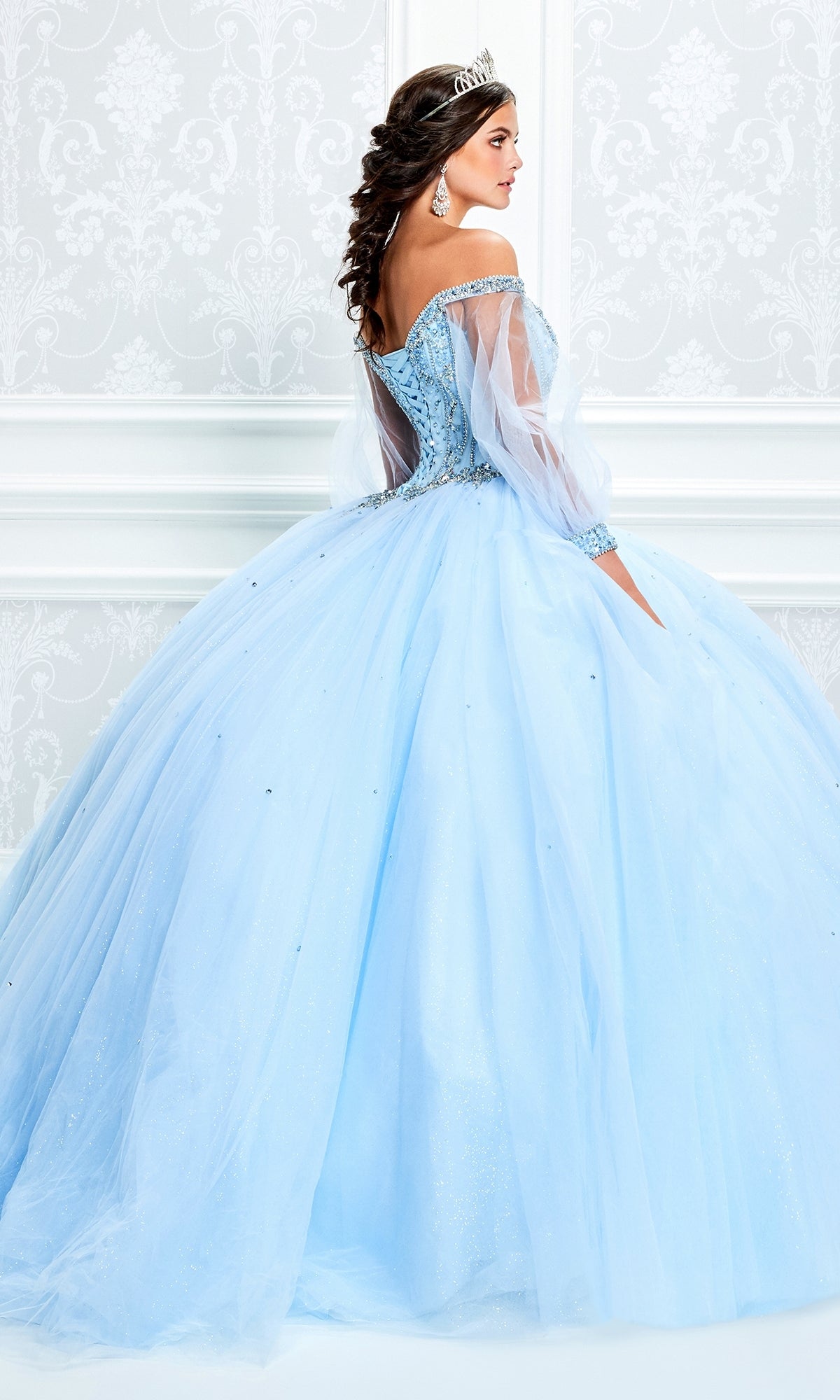 A woman wears the Princesa Light Blue Quinceañera Dress PR11937 with sheer sleeves and a tiara, standing in front of an ornate white wall, turned slightly away from the camera.
