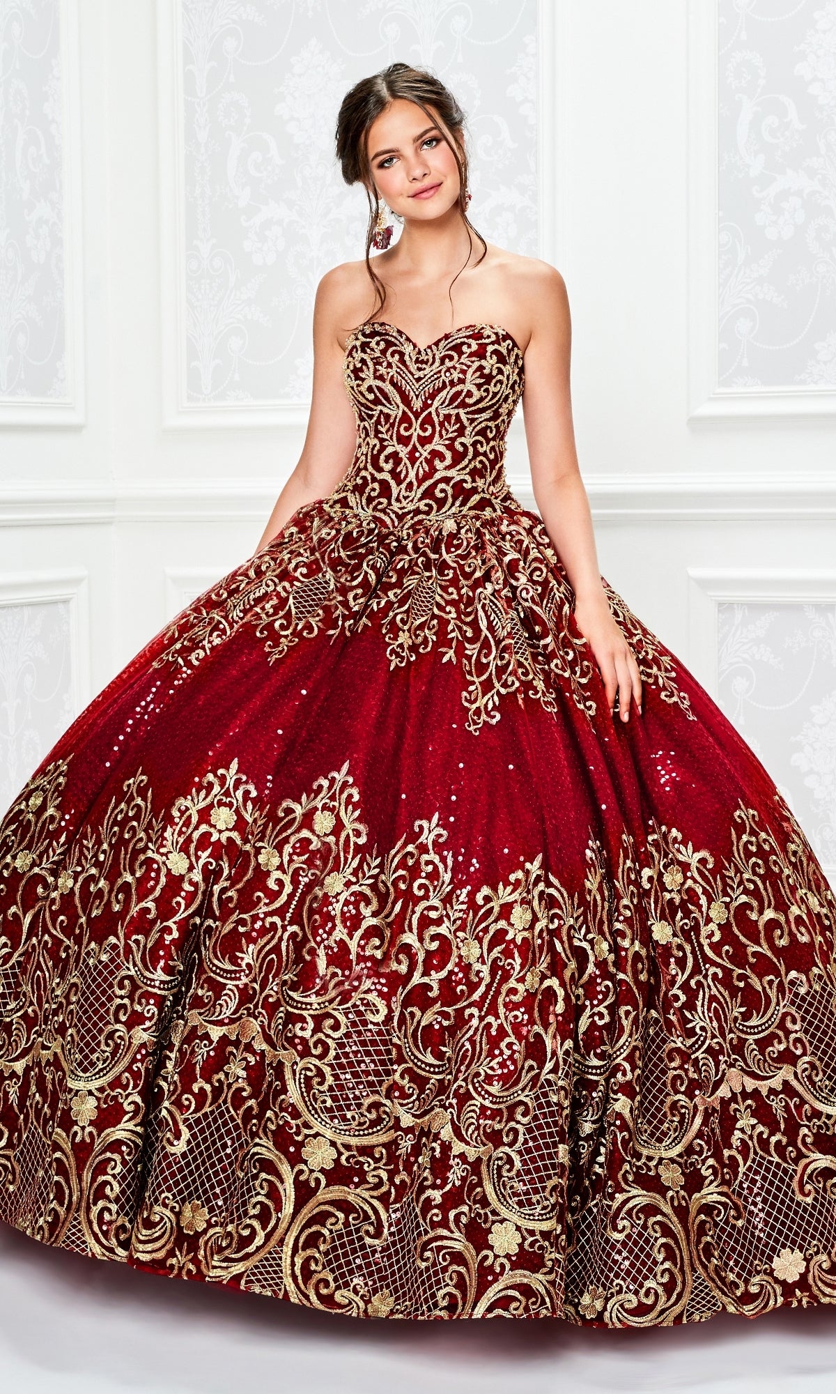 A young woman poses gracefully against a white ornate background, wearing the Princesa Red and Gold Quinceañera Dress PR11945 with elegant gold embroidery.
