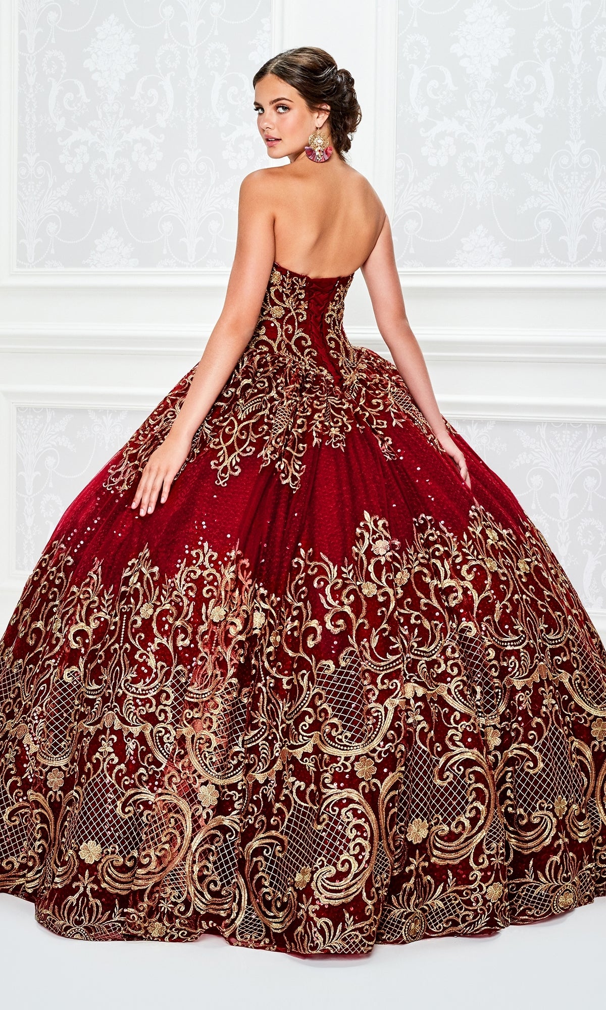 A young woman wearing the Princesa Red and Gold Quinceañera Dress PR11945 stands facing away in an ornate room, looking back over her shoulder.